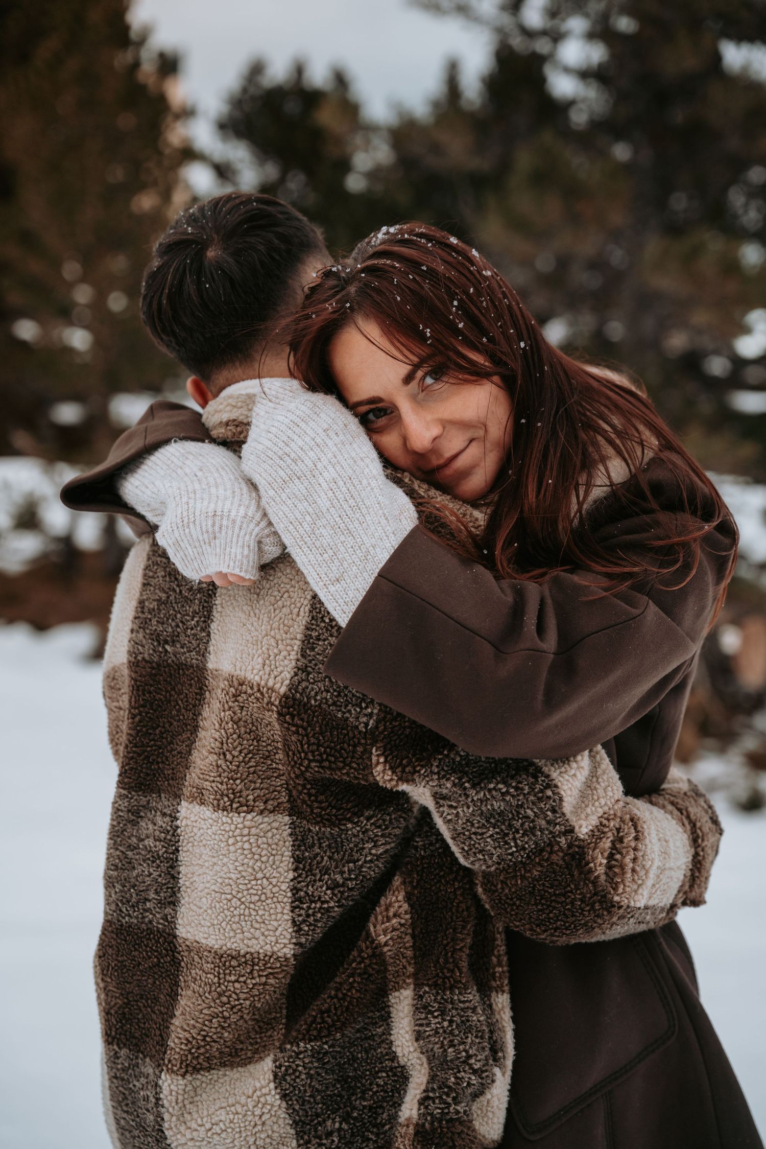 A couple wrapped in a plaid blanket embraces during a winter photoshoot in the snow.