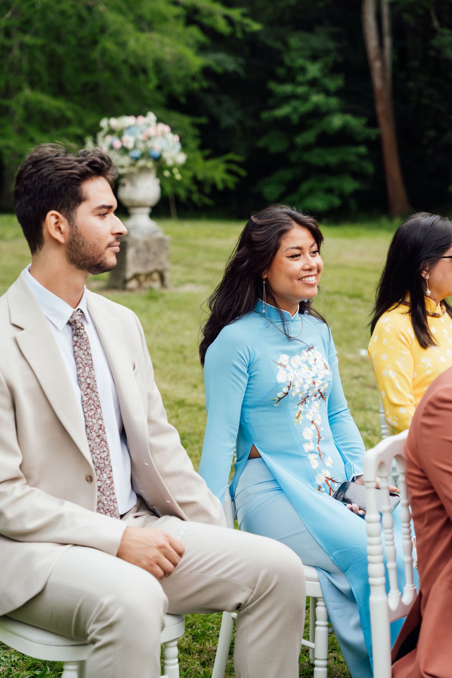 Wedding guests seated outdoors, woman in blue ao dai smiling beside man in beige suit with floral tie.