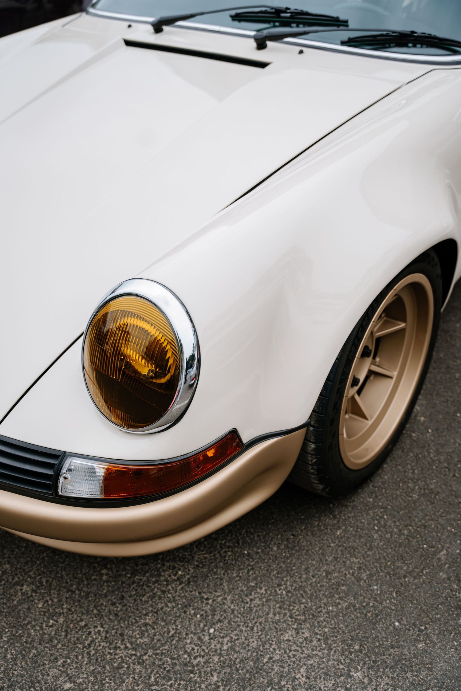 Close-up view of a classic white Porsche 911's front end showing amber headlight and gold wheel detail.