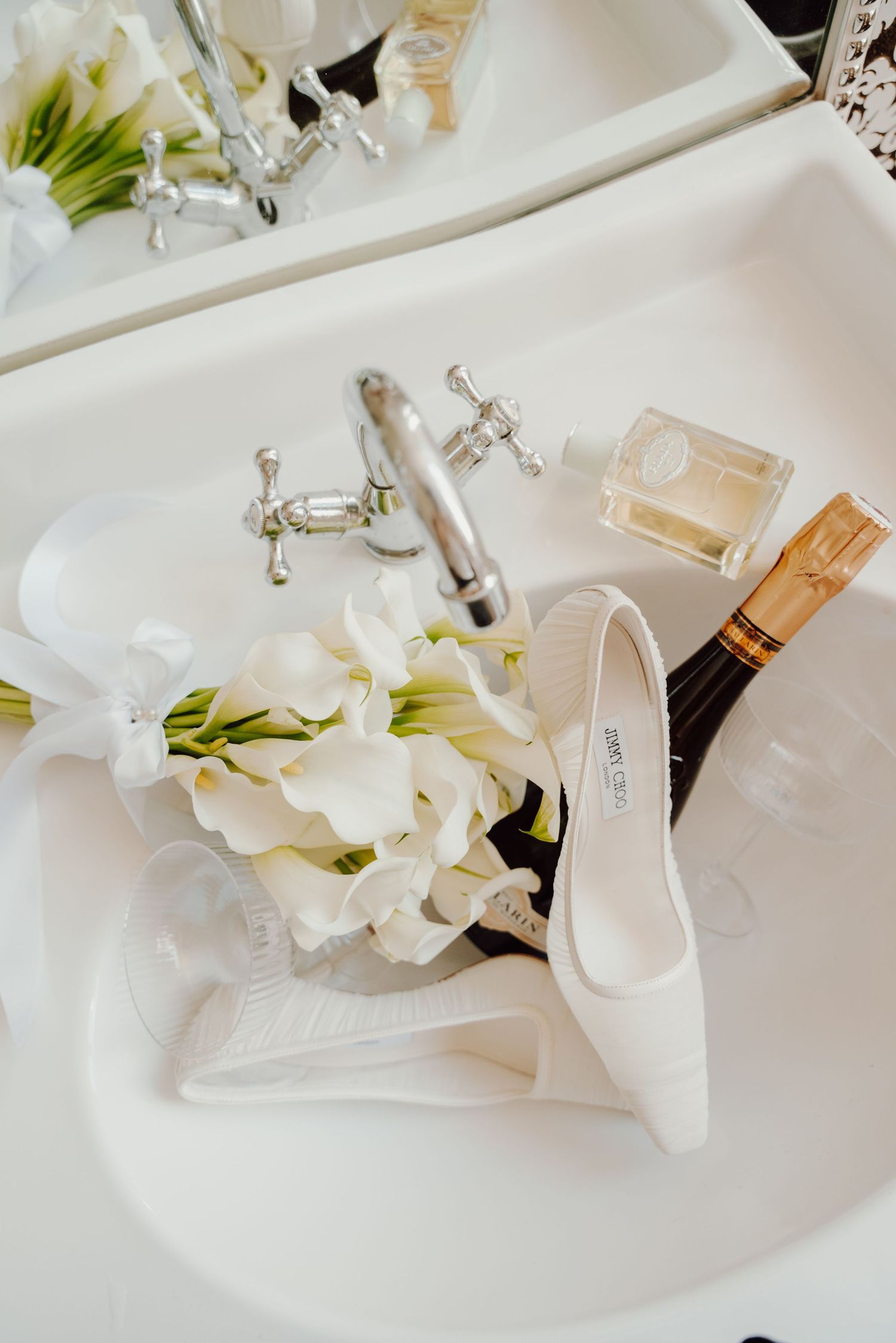Wedding details featuring white calla lilies, perfume, and bridal shoes on white surface.