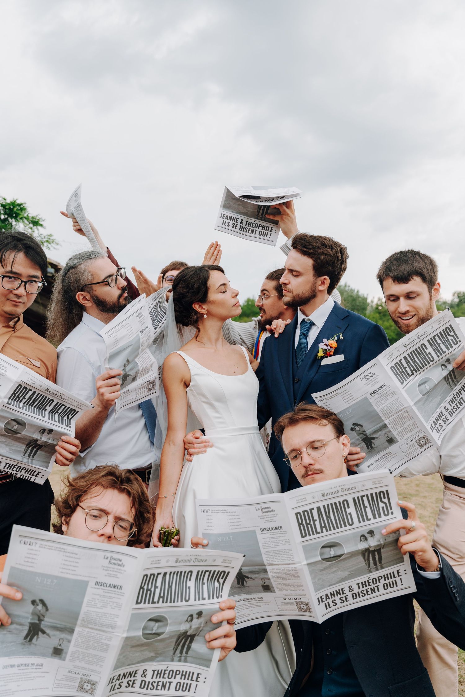 Wedding party poses playfully with newspapers on an overcast day.