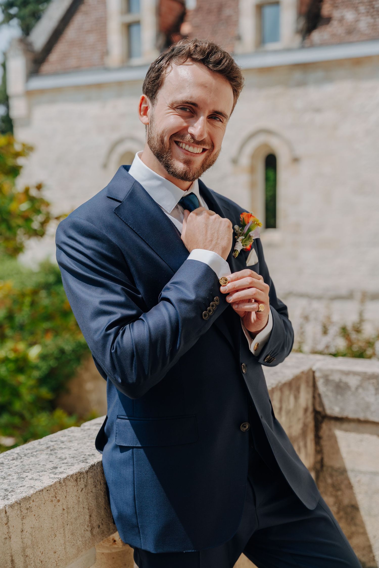 Groom in navy suit adjusts his tie while posing outdoors against stone architecture.