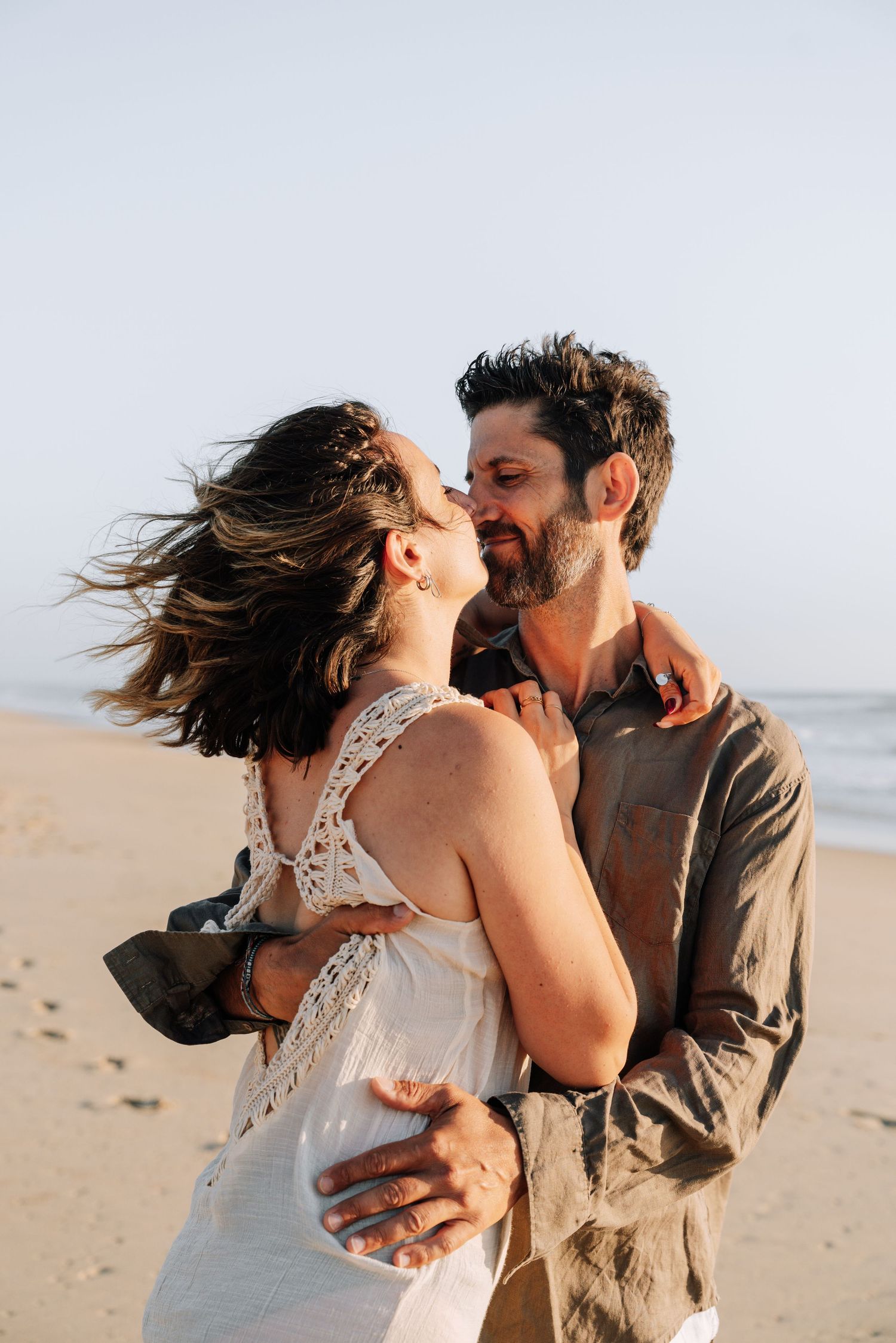 A romantic couple shares tender kisses and embraces while standing on a sunny beach with wind-swept hair.