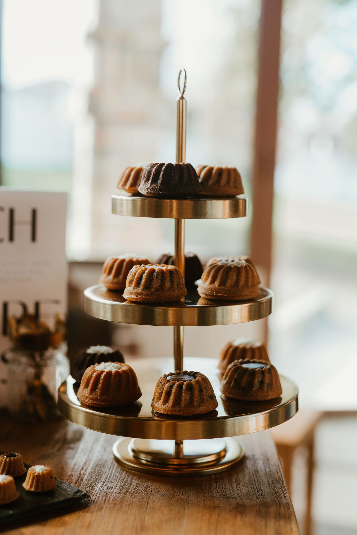Three elegant tiered serving stands display chocolate bundt cakes on metallic gold plates in a bright window-lit setting.