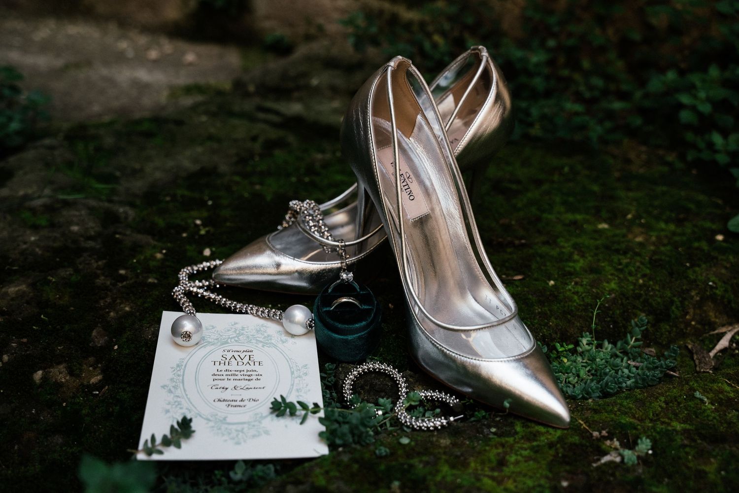 Wedding accessories including silver shoes, jewelry, and invitation details arranged on a dark surface.