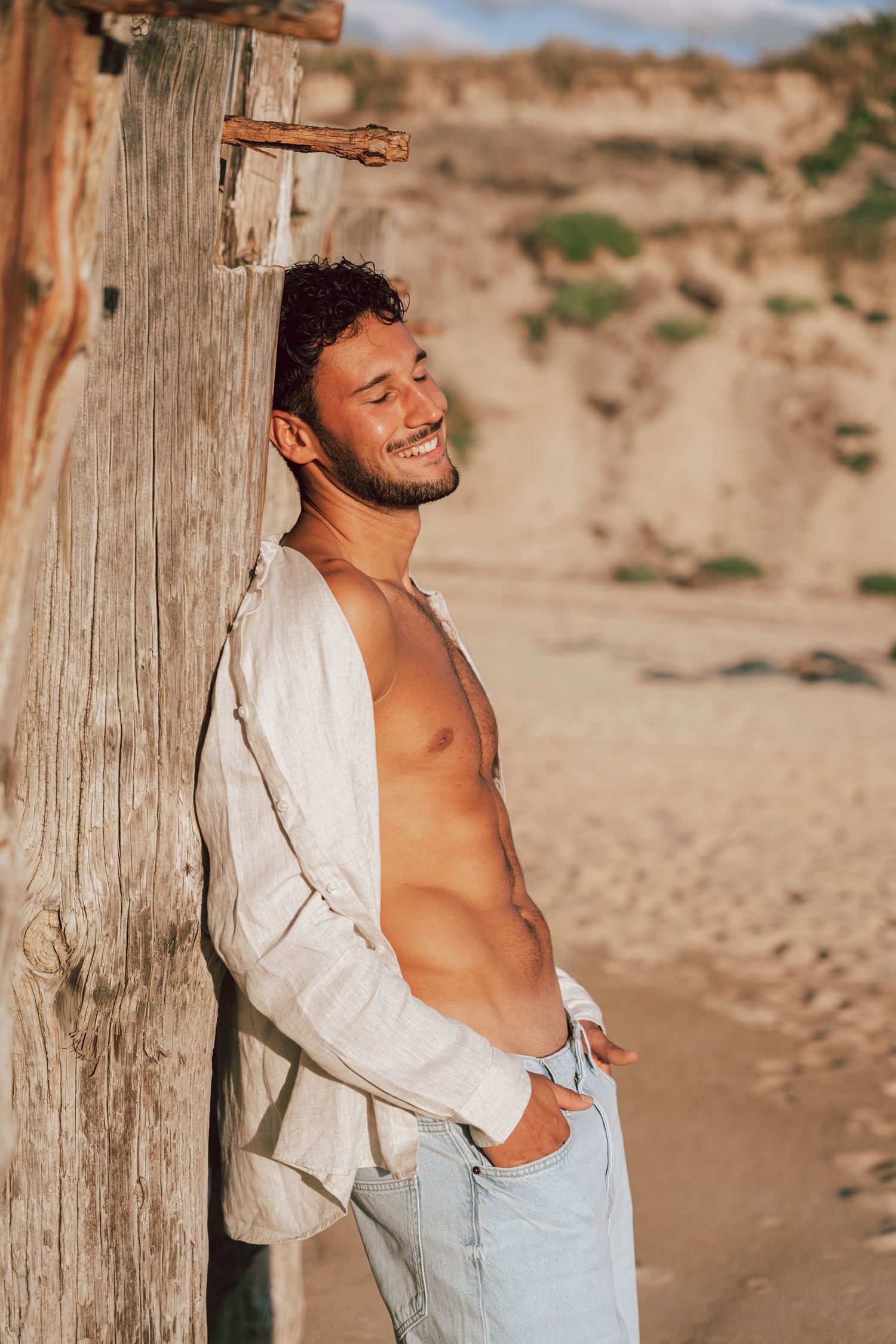 A person in a white shirt laughs joyfully while posing against a wooden post on a sandy beach.