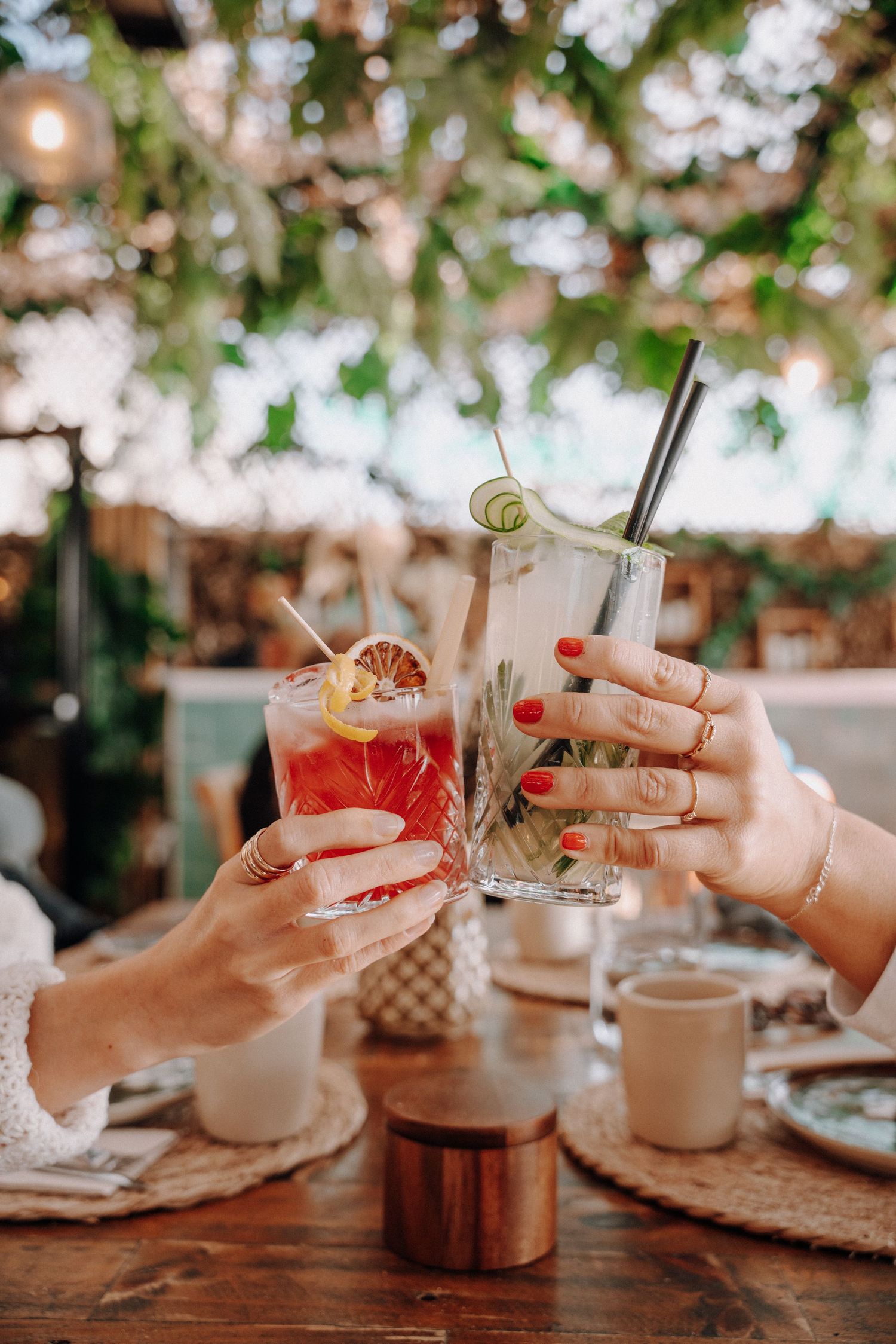 Two hands toasting with colorful cocktails at a rustic outdoor table setting with bokeh lighting in the background.