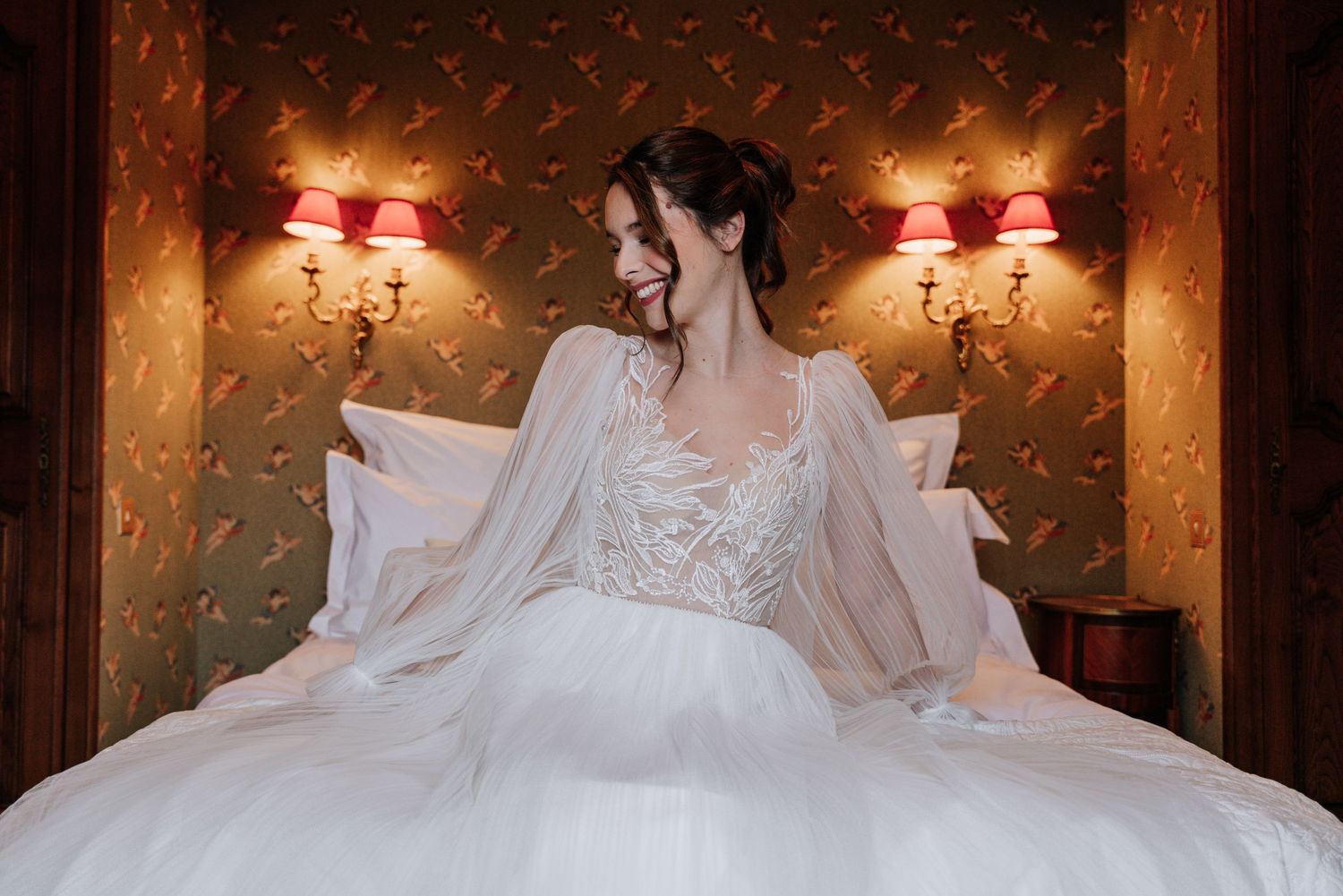 Bride in flowing white dress sitting on luxurious bed with vintage wallpaper background.