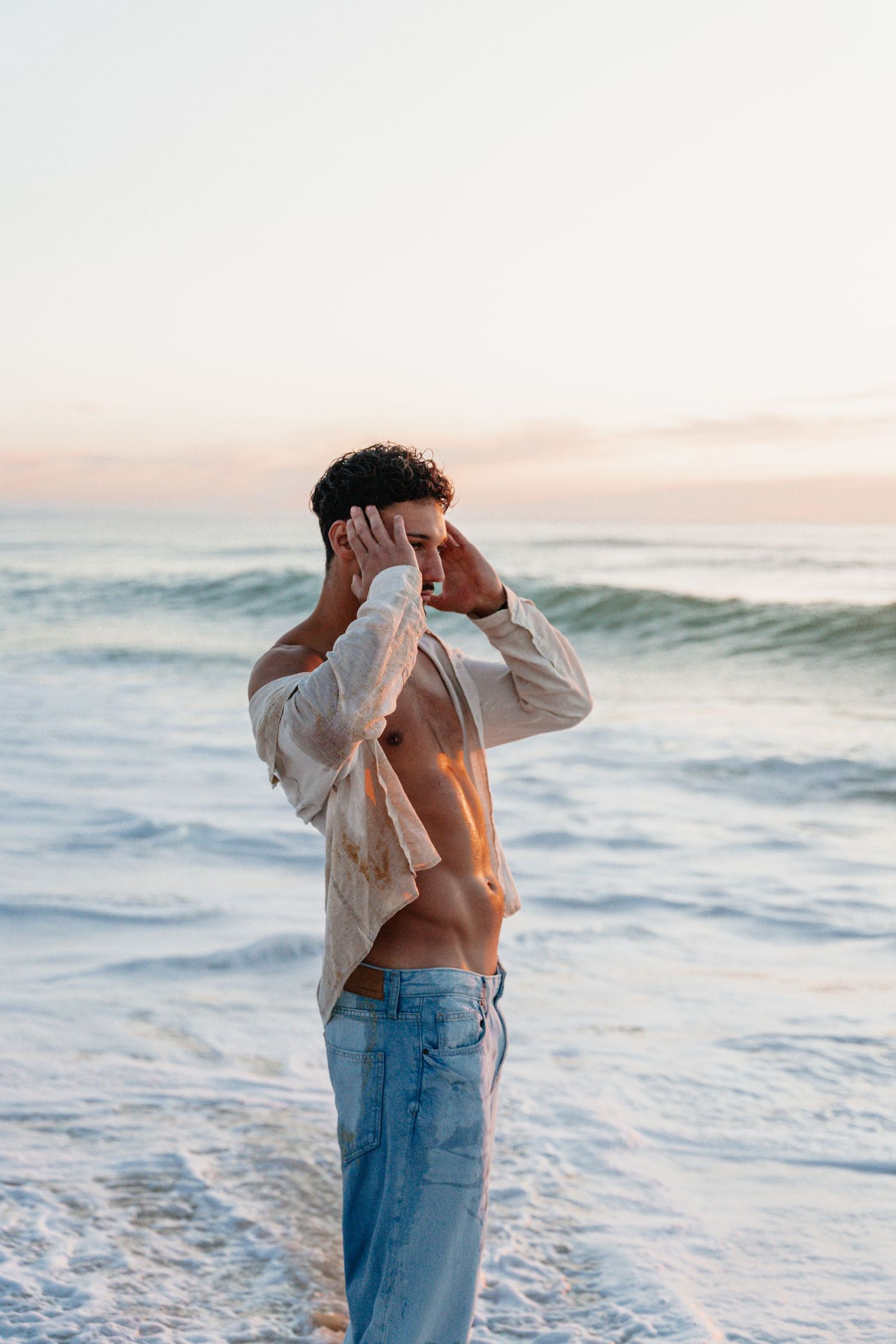 Person in jeans and white shirt poses against ocean waves at sunset in artistic beach photoshoot series.
