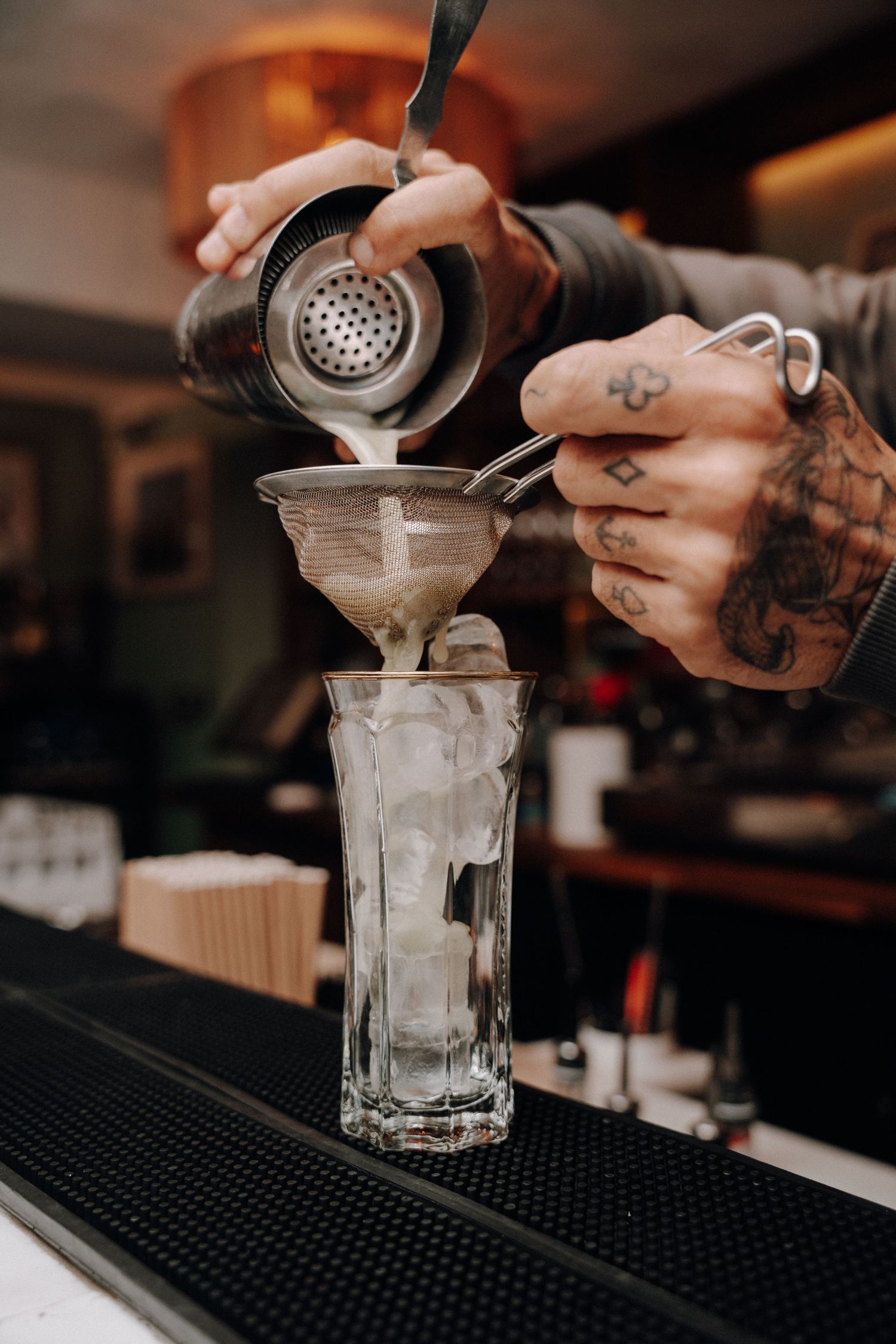 Bartender straining cocktail through Hawthorne strainer into glass with ice at dimly lit bar counter.