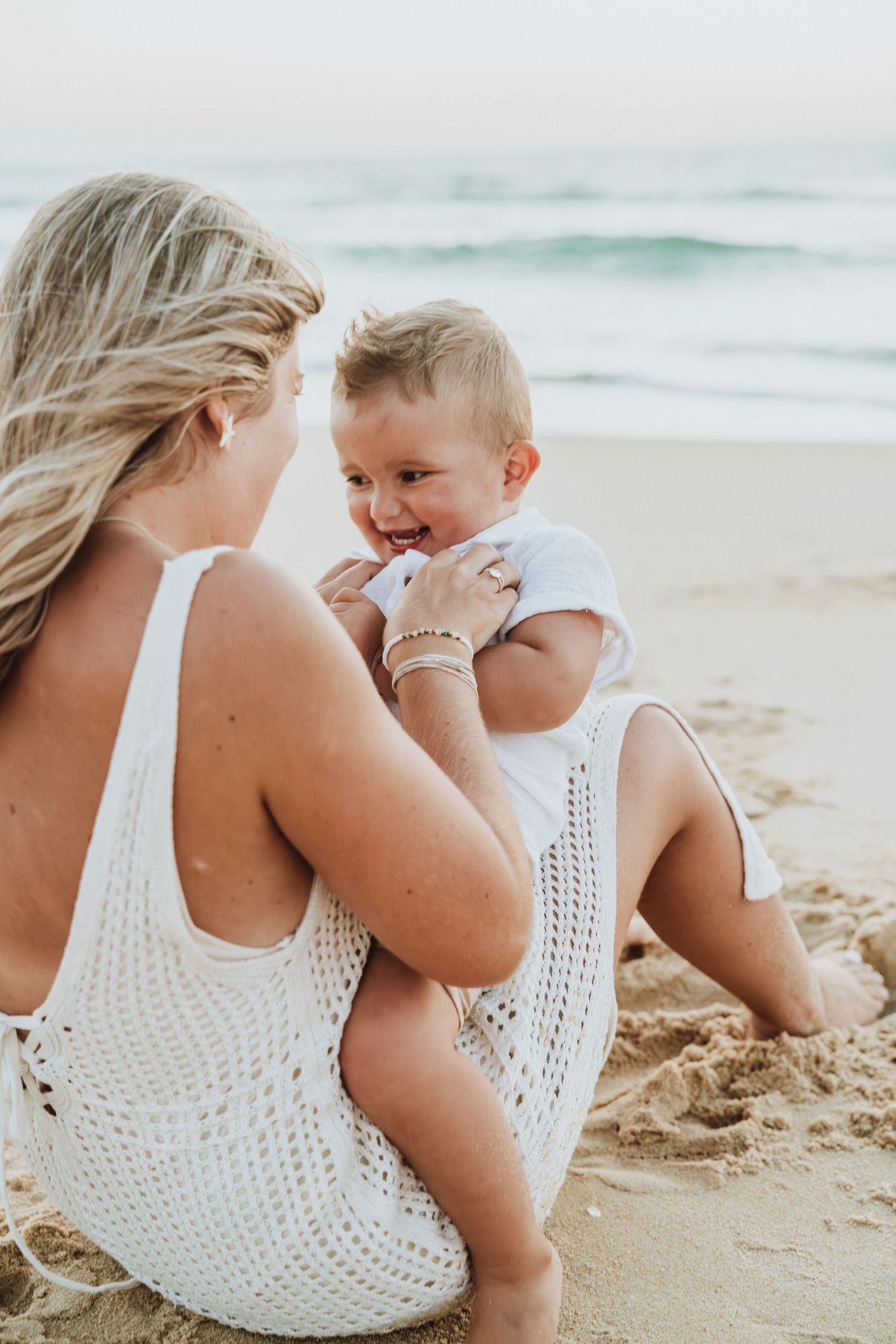 Parent and baby playing together on sandy beach in matching white outfits.
