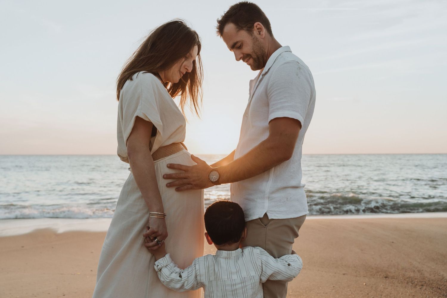 Romantic maternity photoshoot on a beach at sunset with couple embracing and showing pregnant belly.