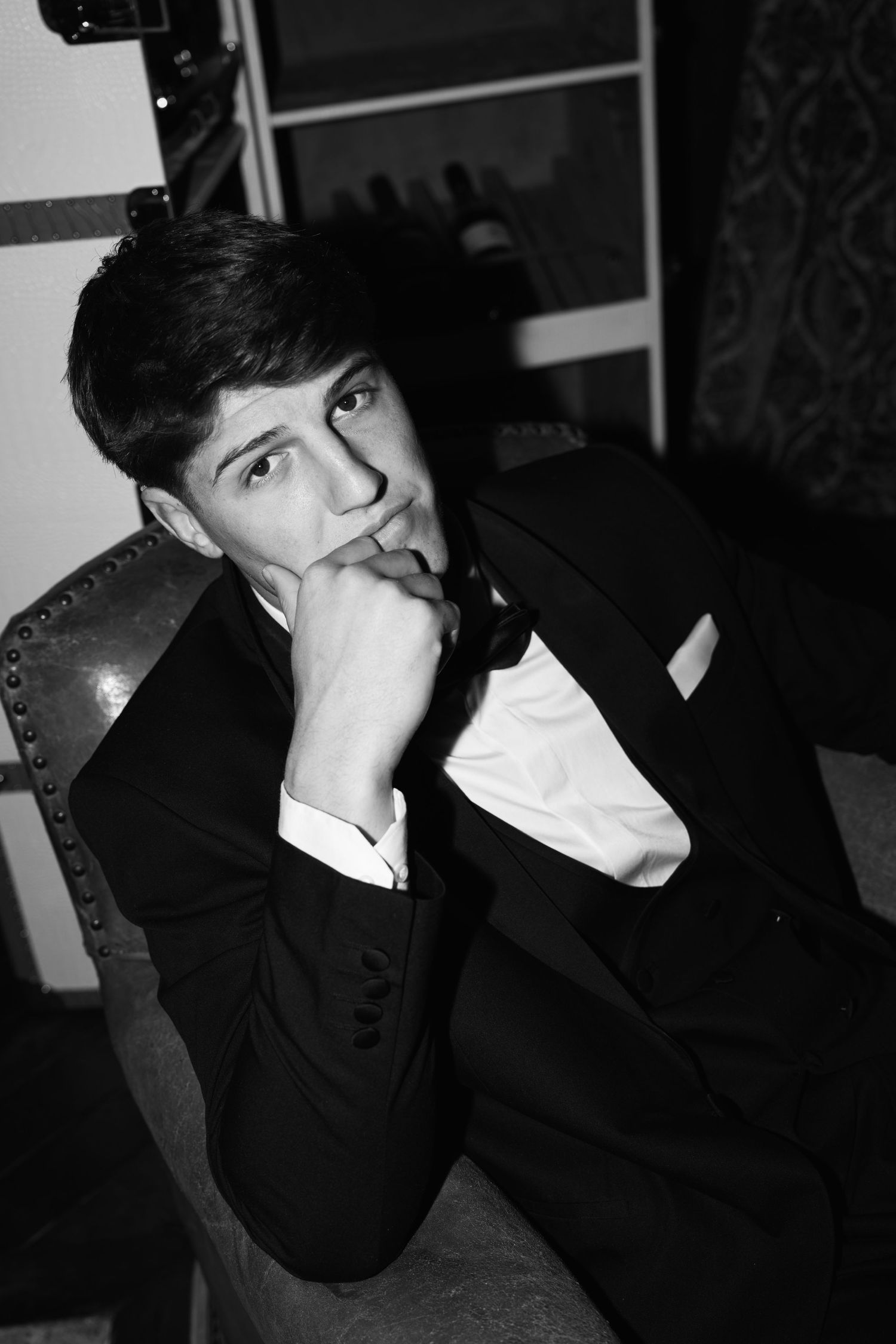 Young man in tuxedo sits pensively with hand near chin, black and white formal portrait photography.