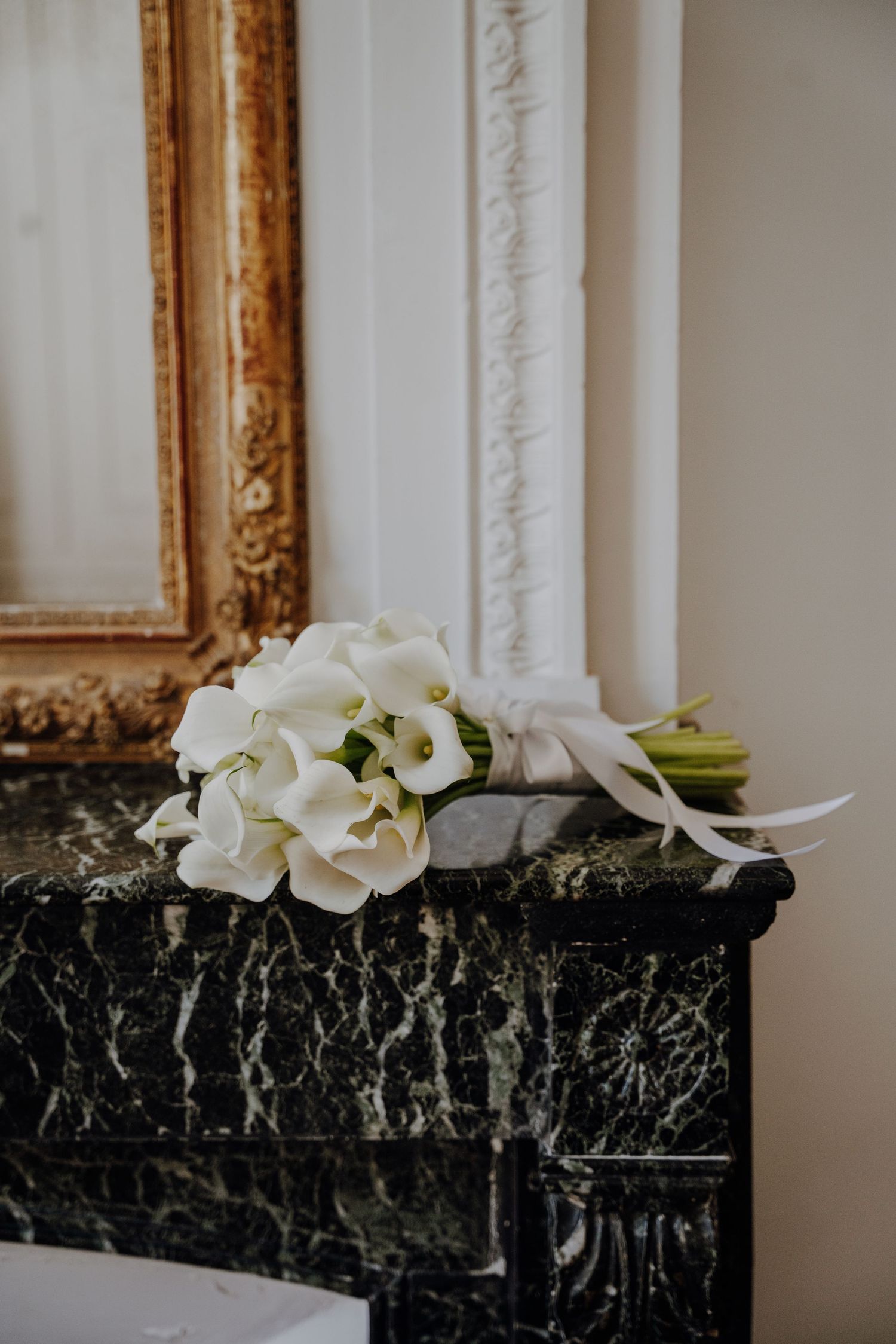 White calla lily bouquet resting on black marble mantelpiece with gold frame.