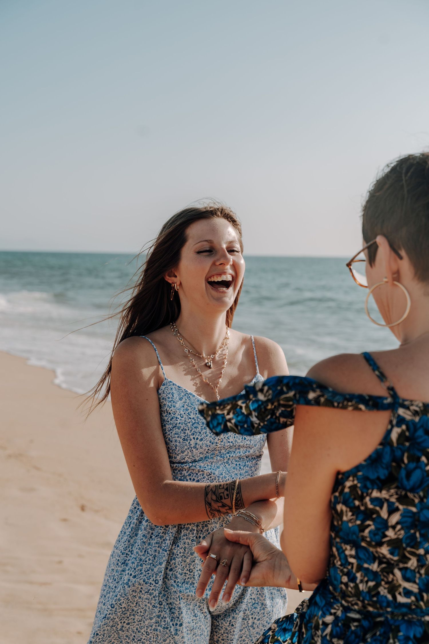 Two people in floral dresses sharing laughter on sunny beach.