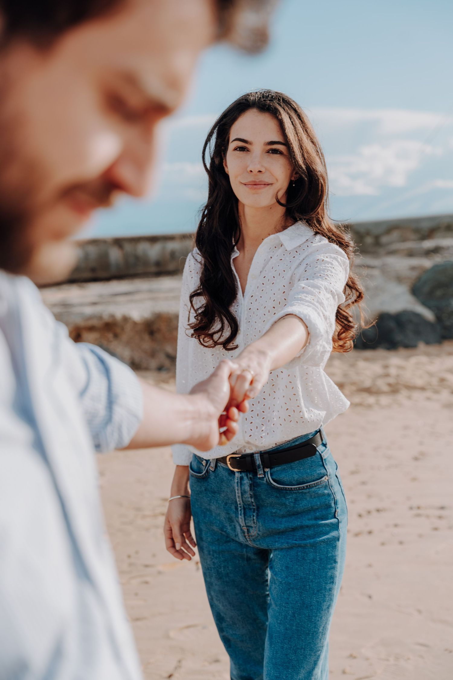 Woman in white top and jeans poses playfully on a beach during a sunny day.