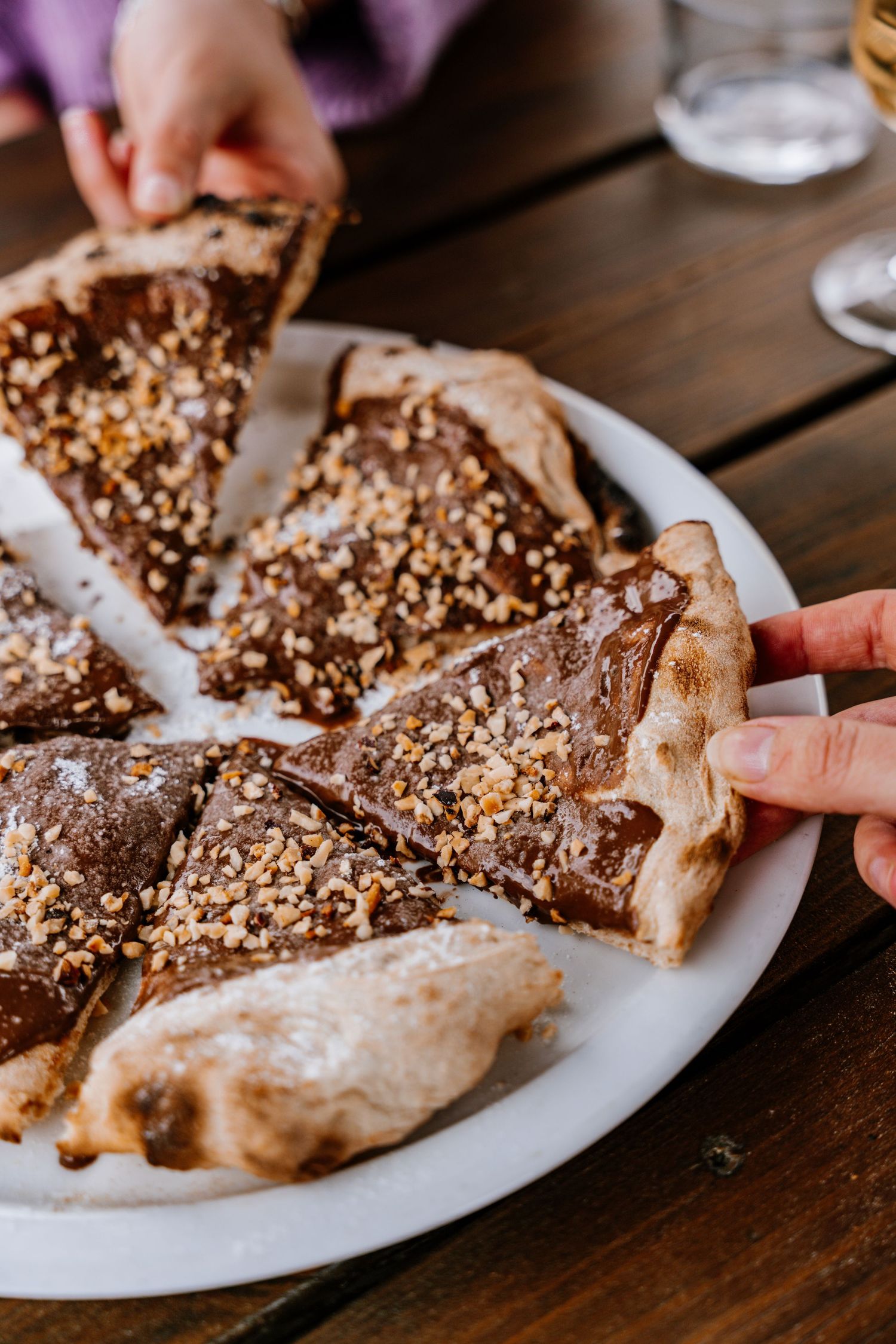 Hands reaching for slices of chocolate-covered flatbread topped with chopped nuts on a white plate.