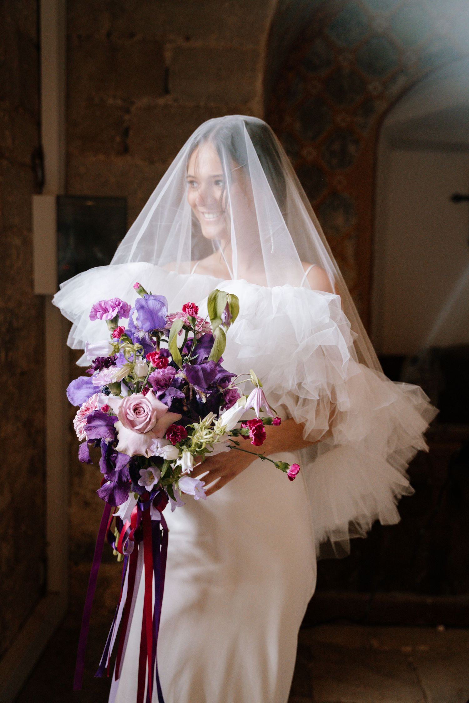 Bridal bouquet with purple and pink flowers against white wedding dress with flowing veil.