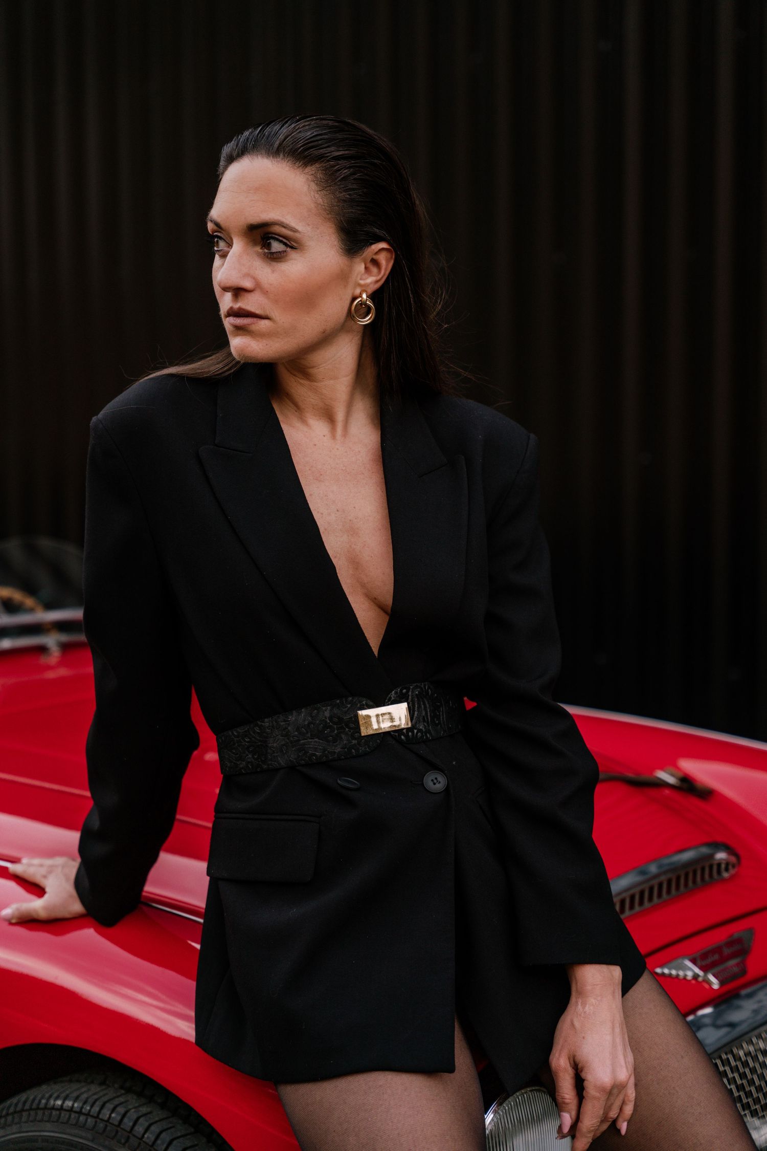 Woman in black blazer posing dramatically near red vintage car with dark background.