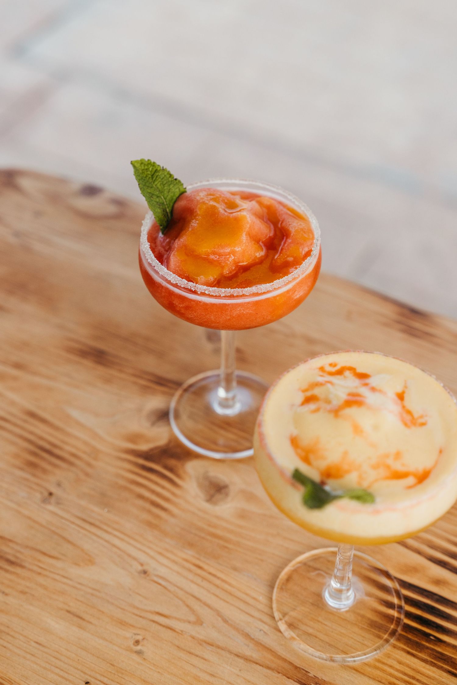 Orange cocktail garnished with mint leaf served in a coupe glass on wooden surface.