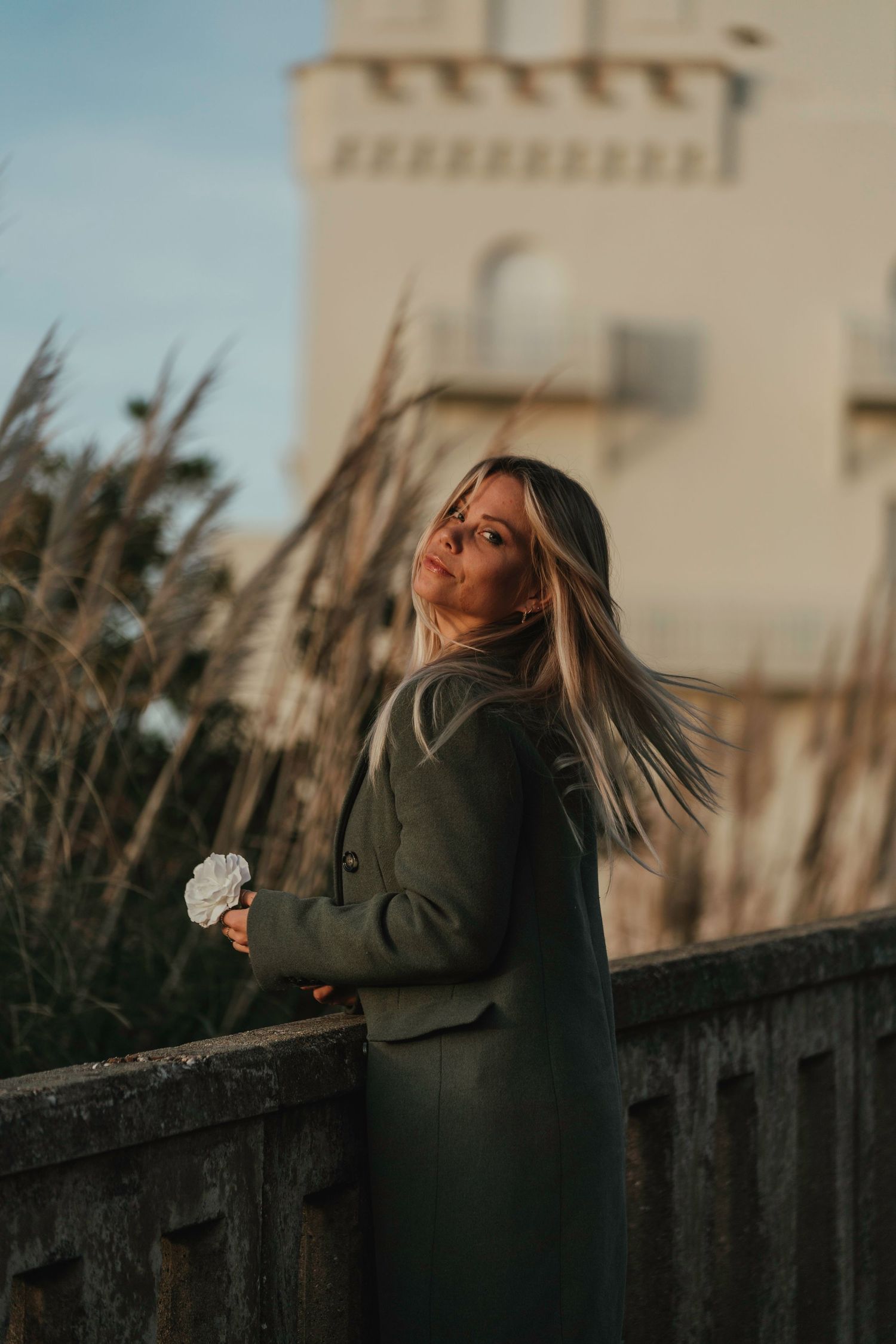 A woman in a dark coat stands against a fence at sunset, tossing her blonde hair in dramatic motion.