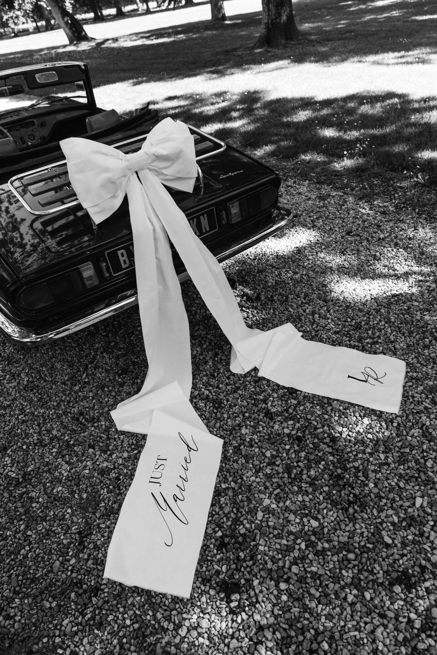 Black and white photo of Just Married ribbon bow on vintage convertible car at wedding reception.