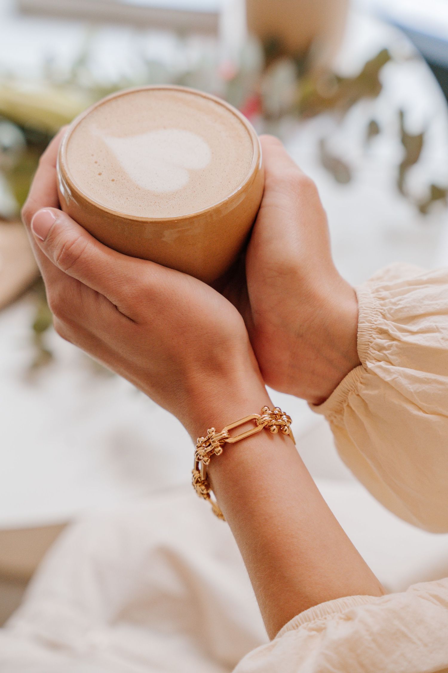 Hands holding a ceramic mug of latte with heart-shaped foam art, wearing a delicate gold charm bracelet and cream sleeve.