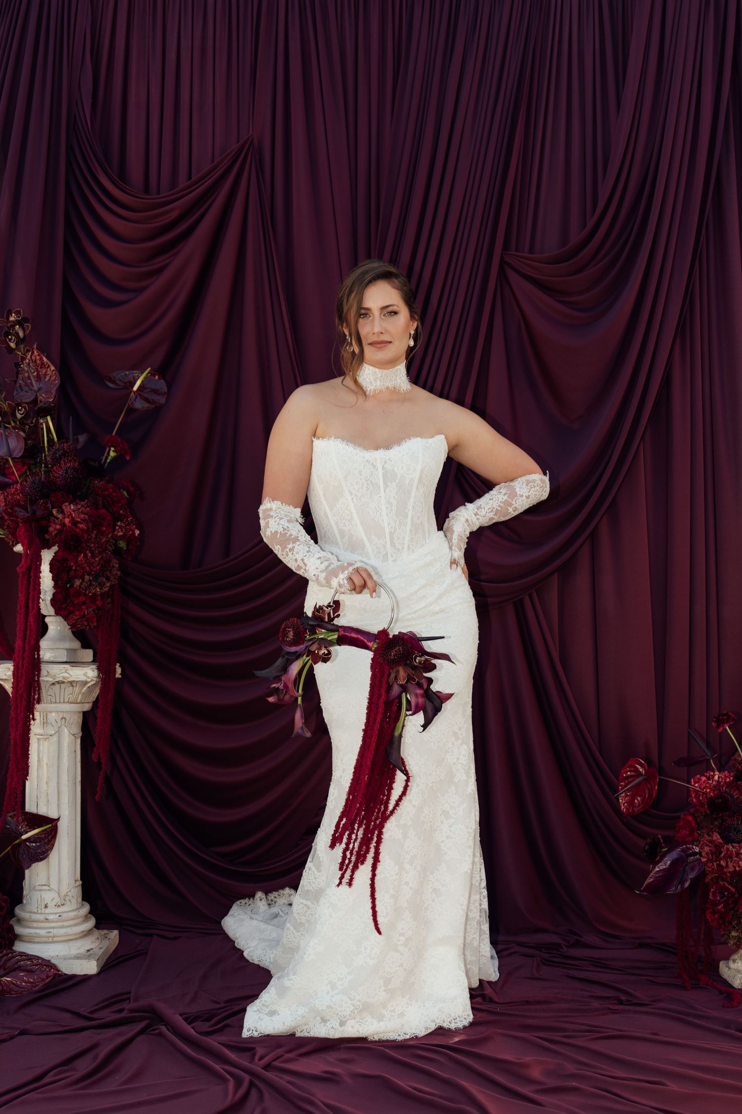 Bride in strapless lace mermaid gown with gloves holding burgundy bouquet against draped purple backdrop.