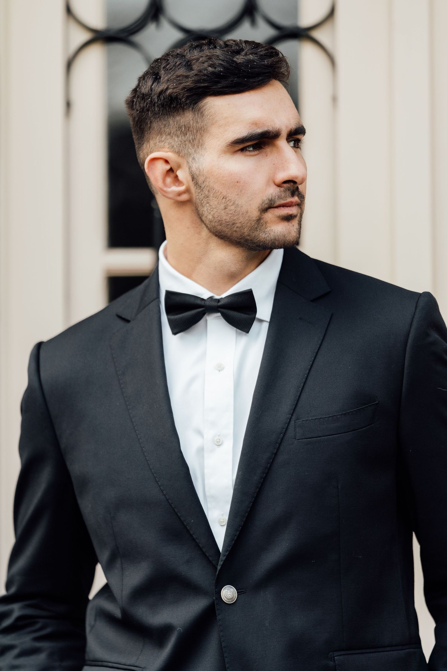A well-dressed man in a black tuxedo with bow tie gazes confidently to the side outdoors.