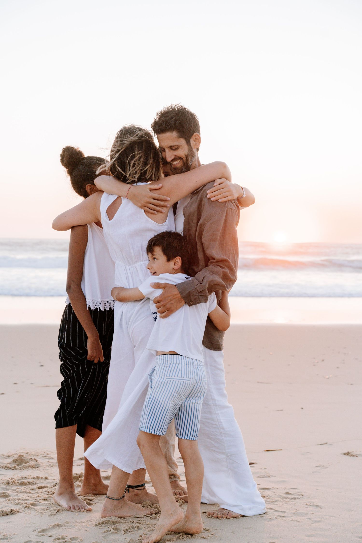 Family embracing together on a sandy beach at sunset in white and casual summer clothing.
