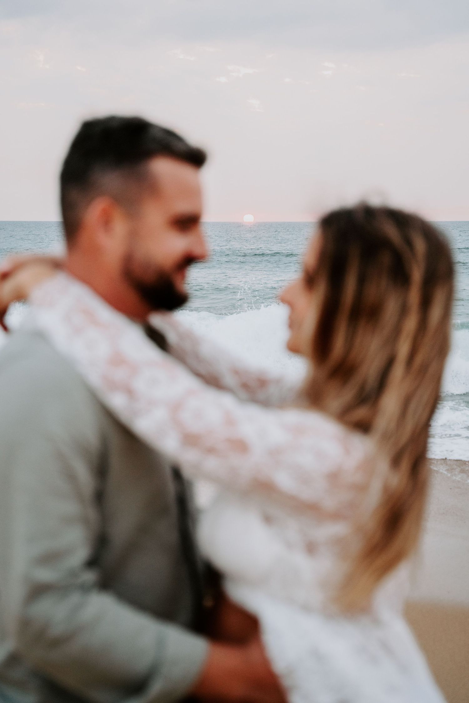 A romantic beach wedding photoshoot showing a couple in white and gray attire dancing by the ocean at sunset.