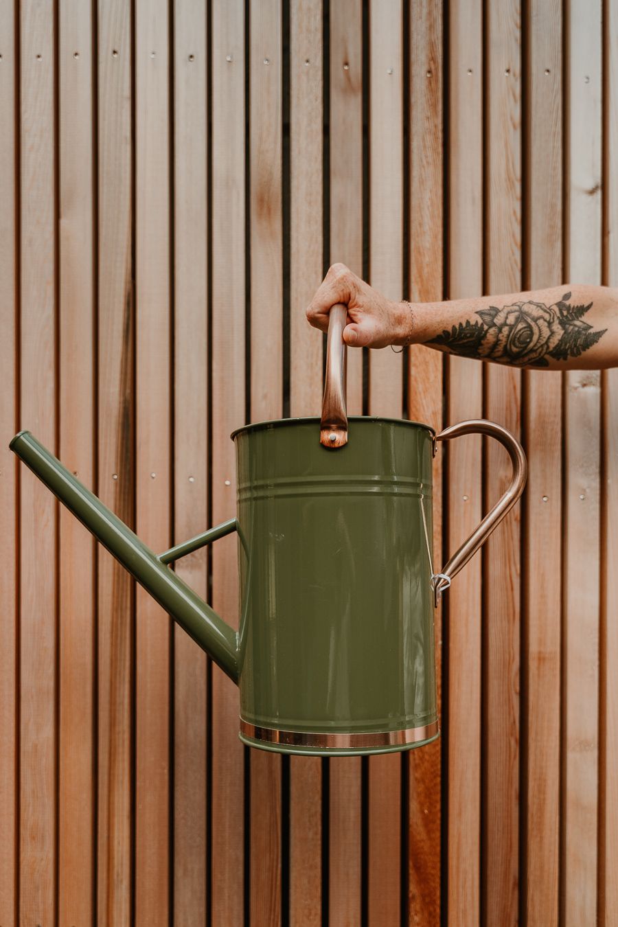 A tattooed arm holds a vintage-style olive green metal watering can against wooden slat background.