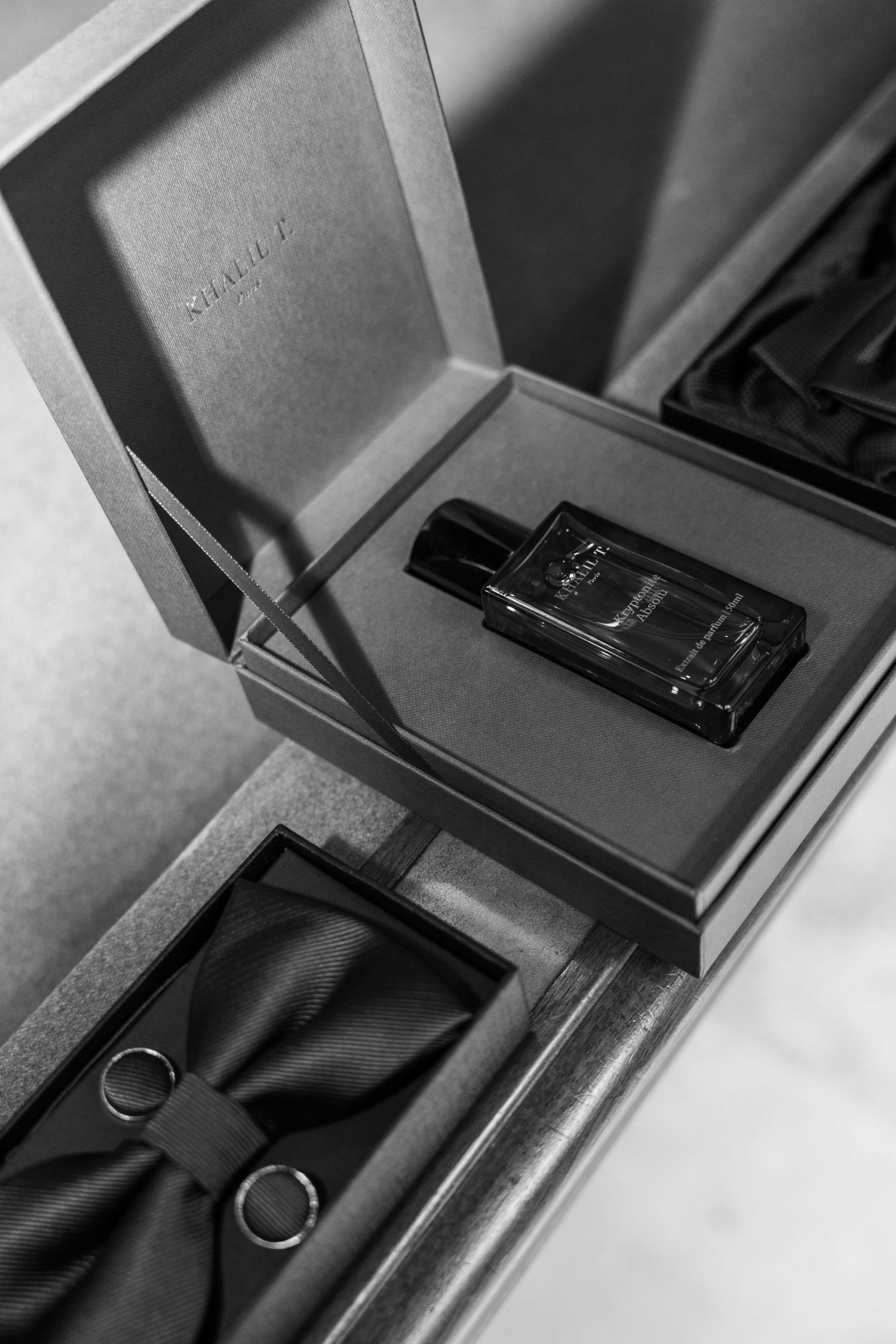 Black and white photo of luxury wedding groom accessories including cologne, bow tie, and rings in elegant gift box.