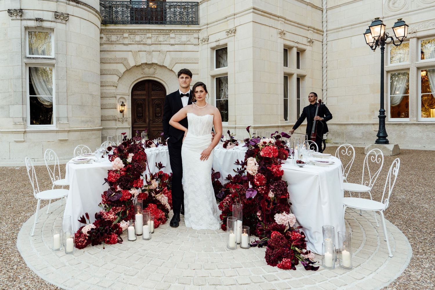 Elegant wedding couple poses before a luxurious floral table display with red blooms and candles at a grand estate.