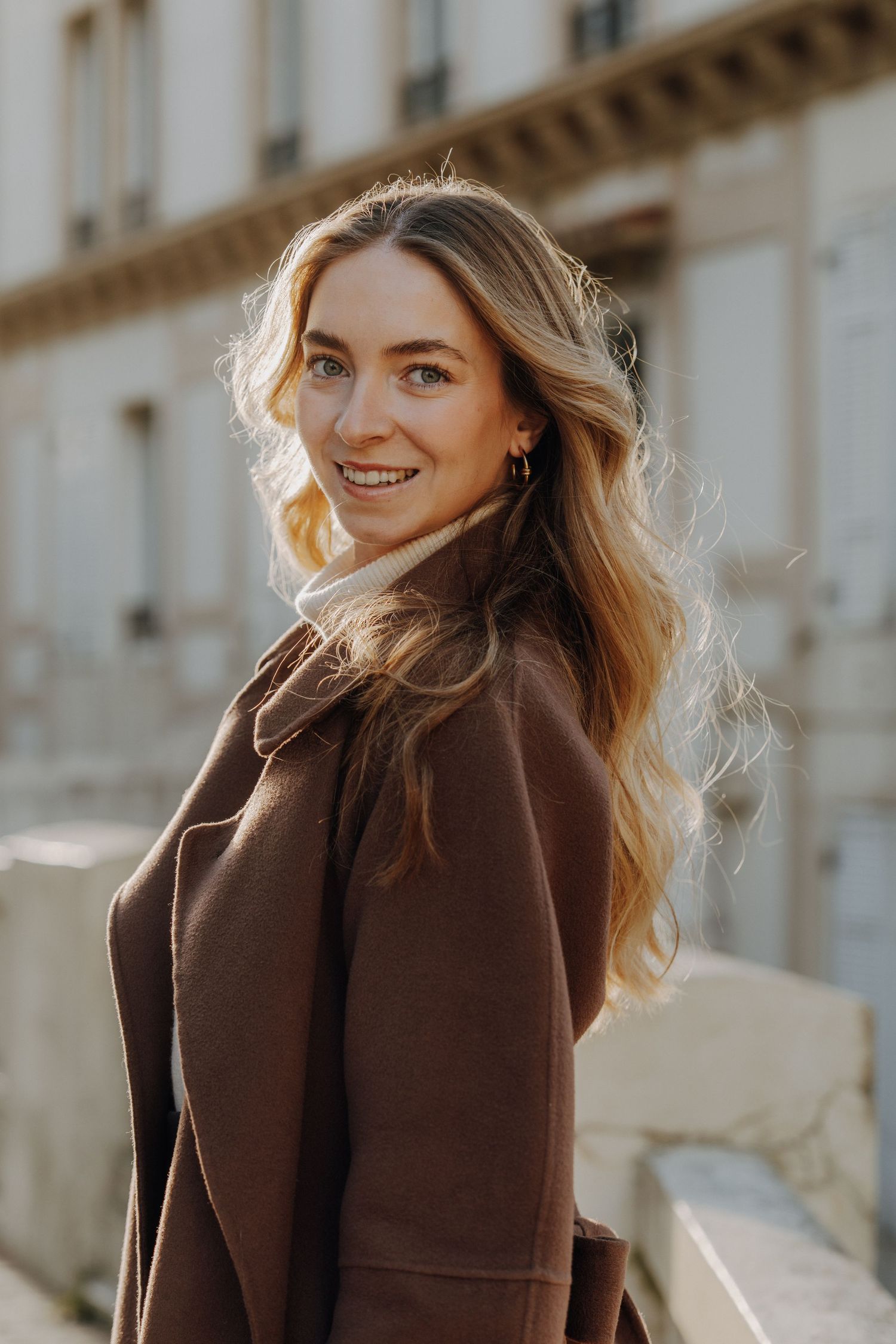 Woman in brown coat smiling with wind-swept hair against architectural backdrop.