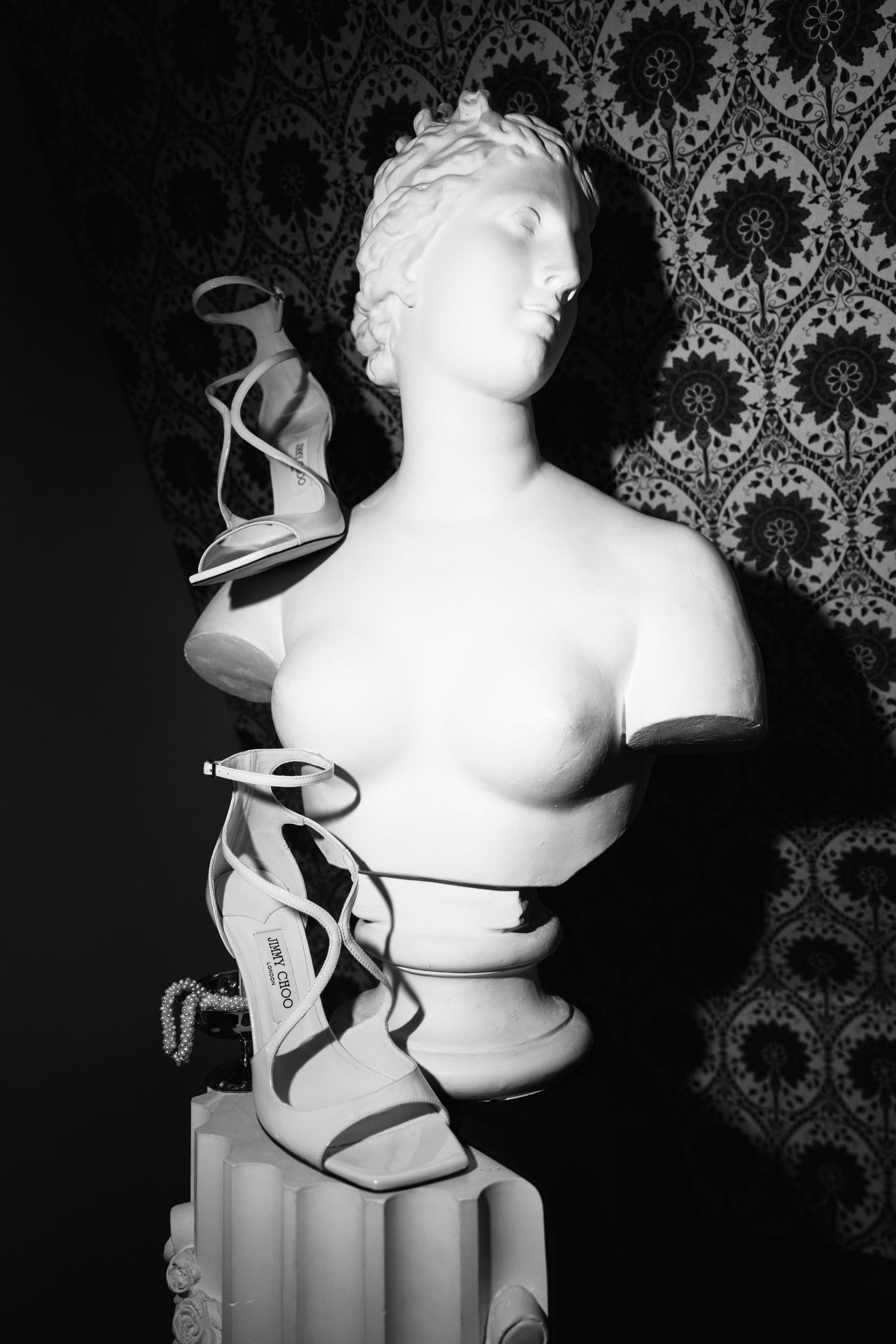 Black and white photo of a classical female bust sculpture with Jimmy Choo strappy heels draped around it.