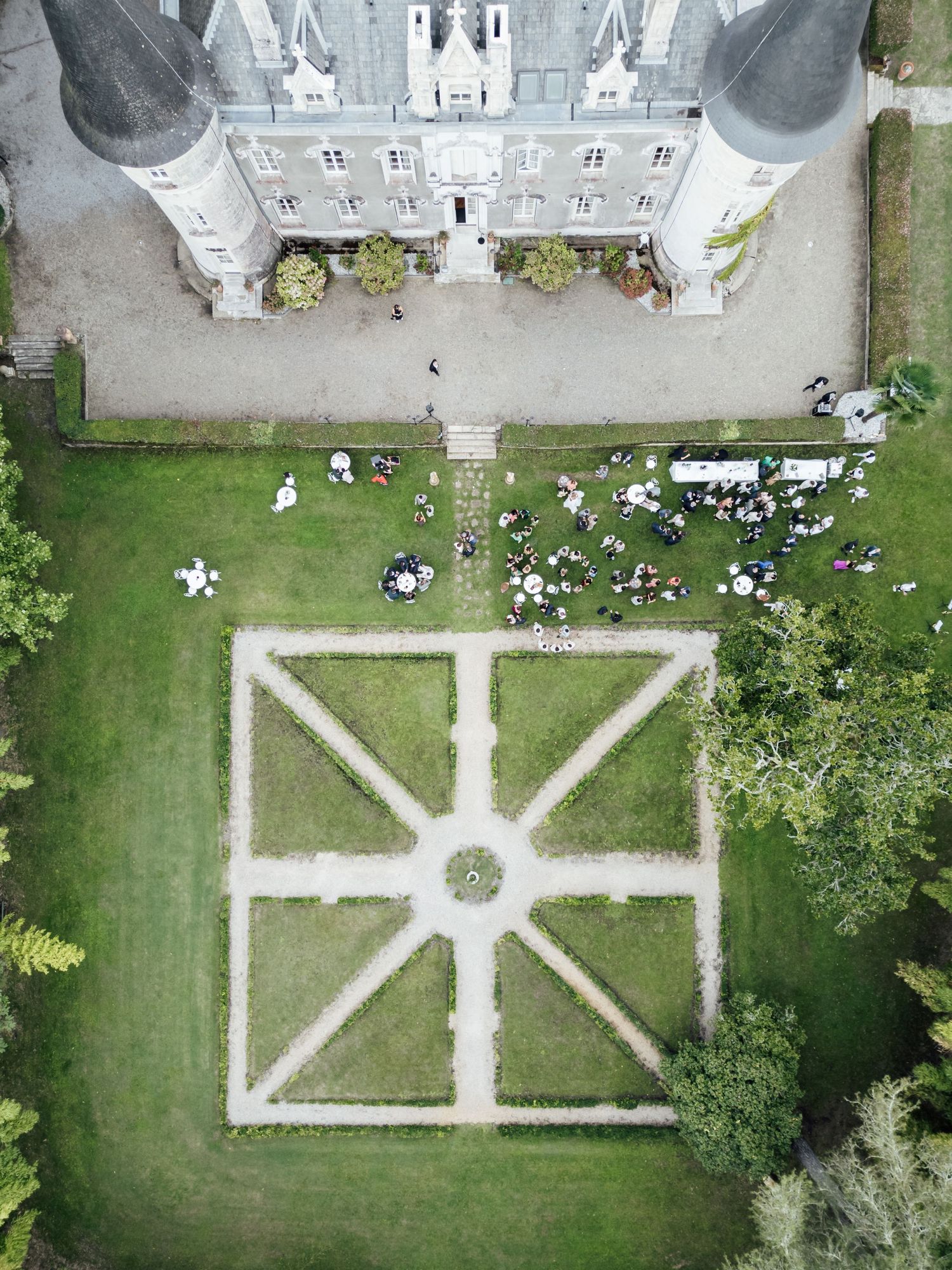 Aerial drone view of a French château with formal geometric garden patterns and wedding guests gathered on the lawn.