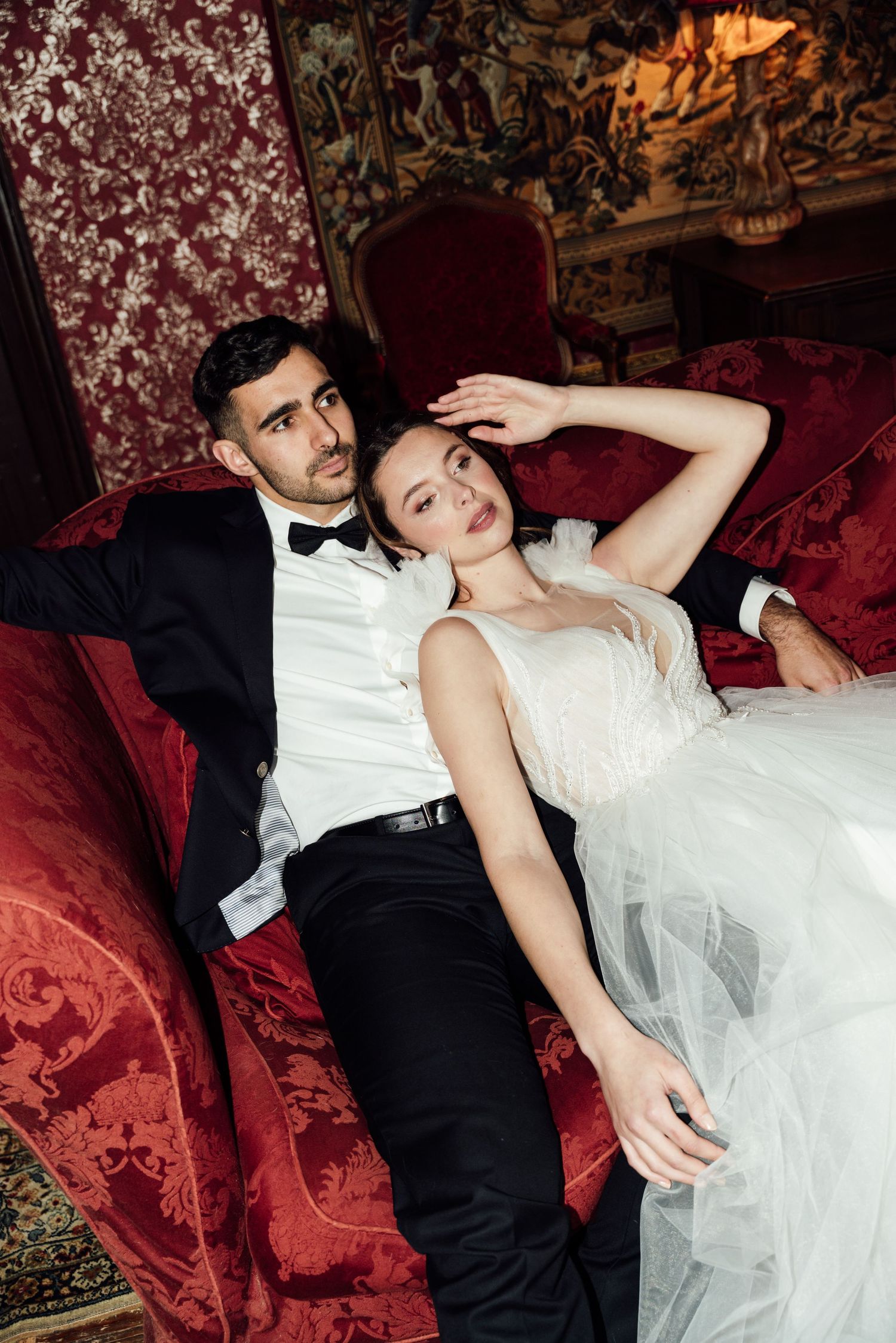 Romantic wedding couple in formal attire lounges on red velvet sofa in vintage-style setting.