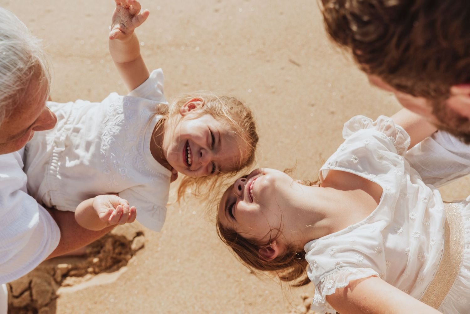 Family playing and laughing together on sandy beach in white outfits.