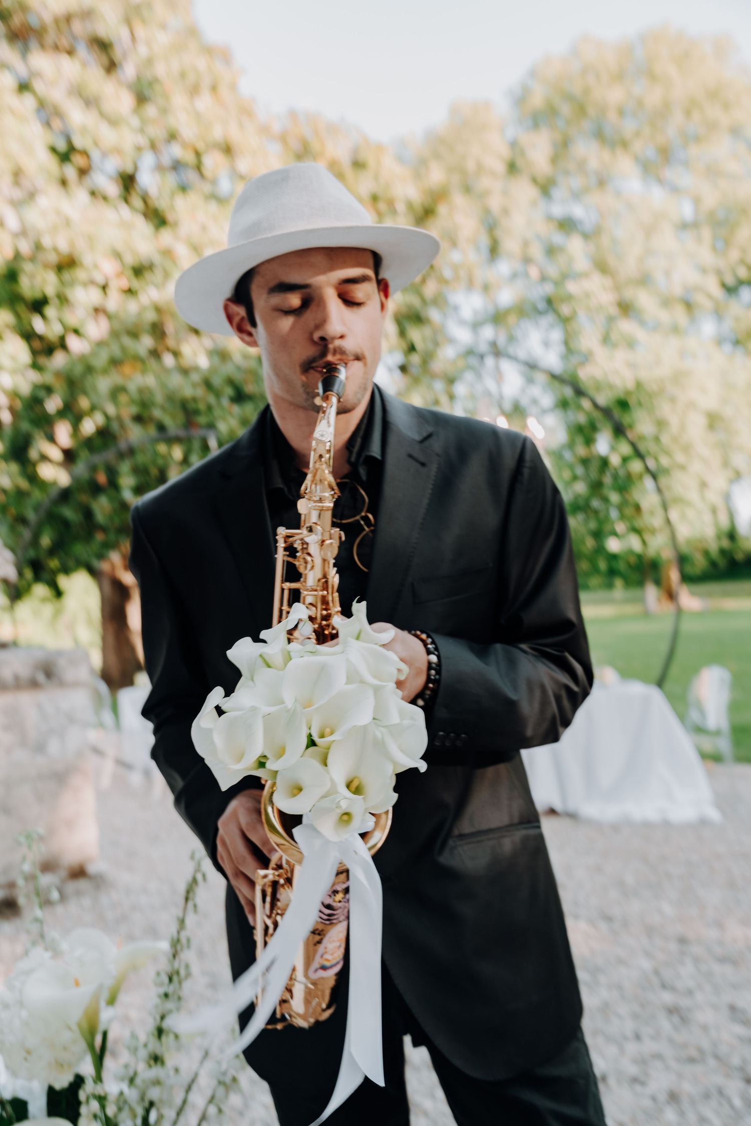 Musician in black suit plays saxophone decorated with white flowers at outdoor event.