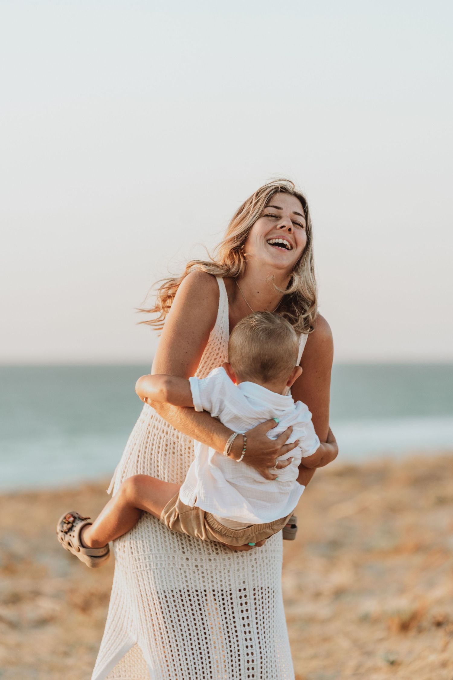Mother and baby sharing joyful laughter while sitting on beach in white crochet dress at sunset.