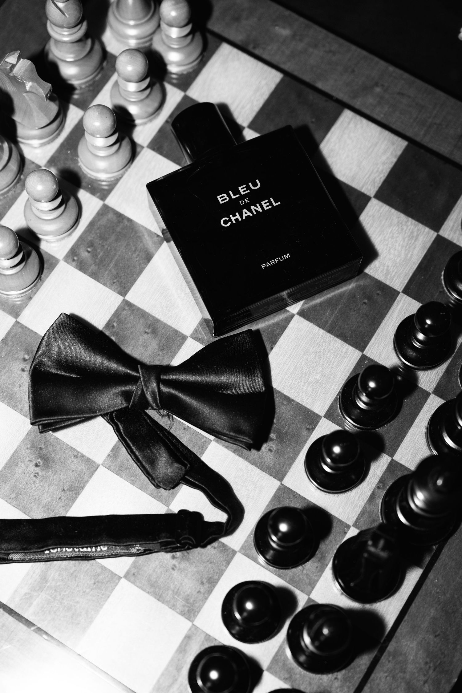 Black and white flat lay of Bleu de Chanel parfum bottle, bow tie, and chess pieces on a chessboard.