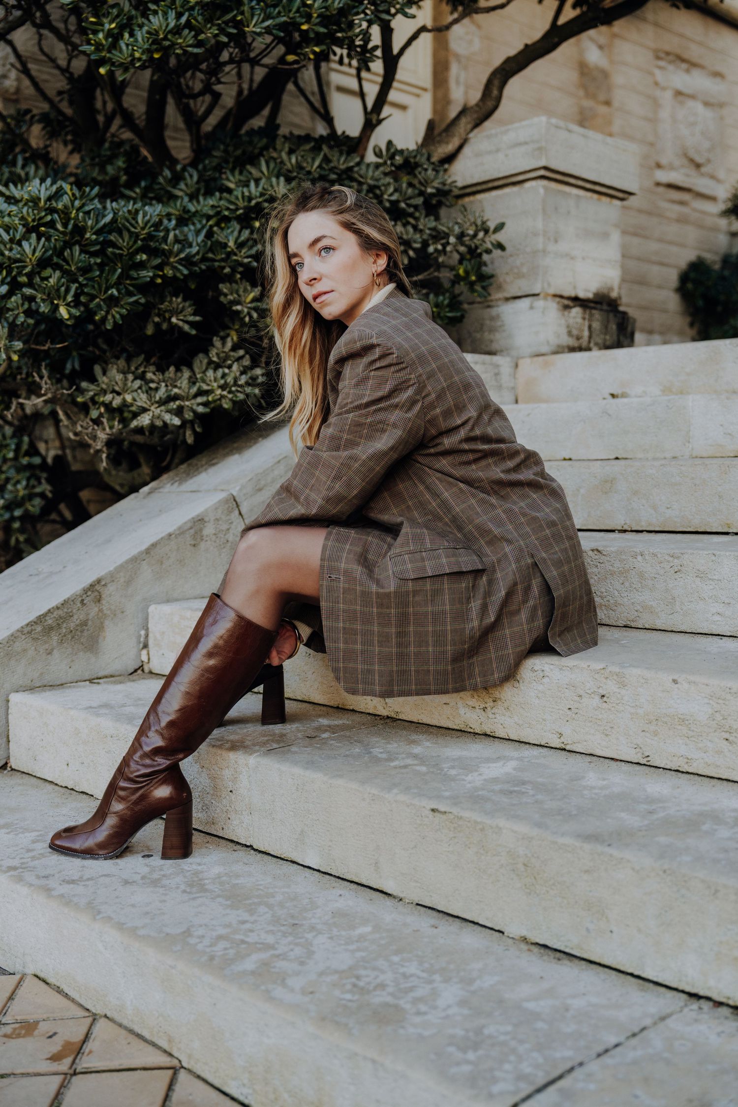 Model wearing plaid blazer and brown boots poses on concrete steps in urban setting.