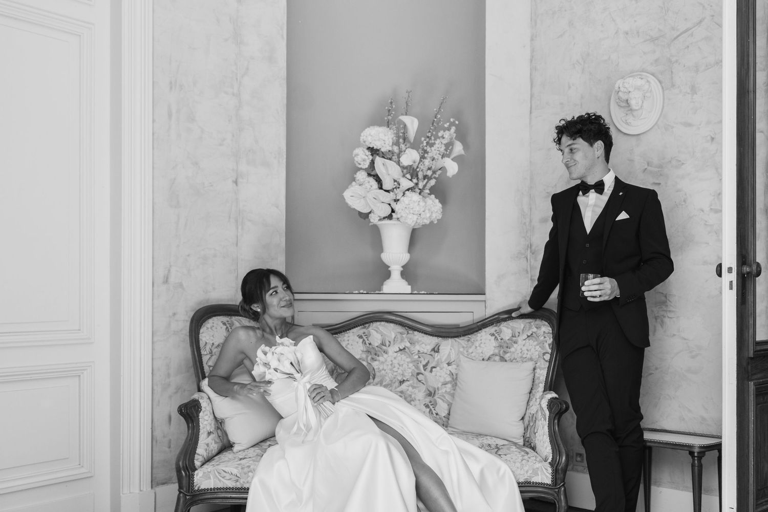 Black and white wedding photo of bride on ornate sofa and groom standing beside her in elegant room.