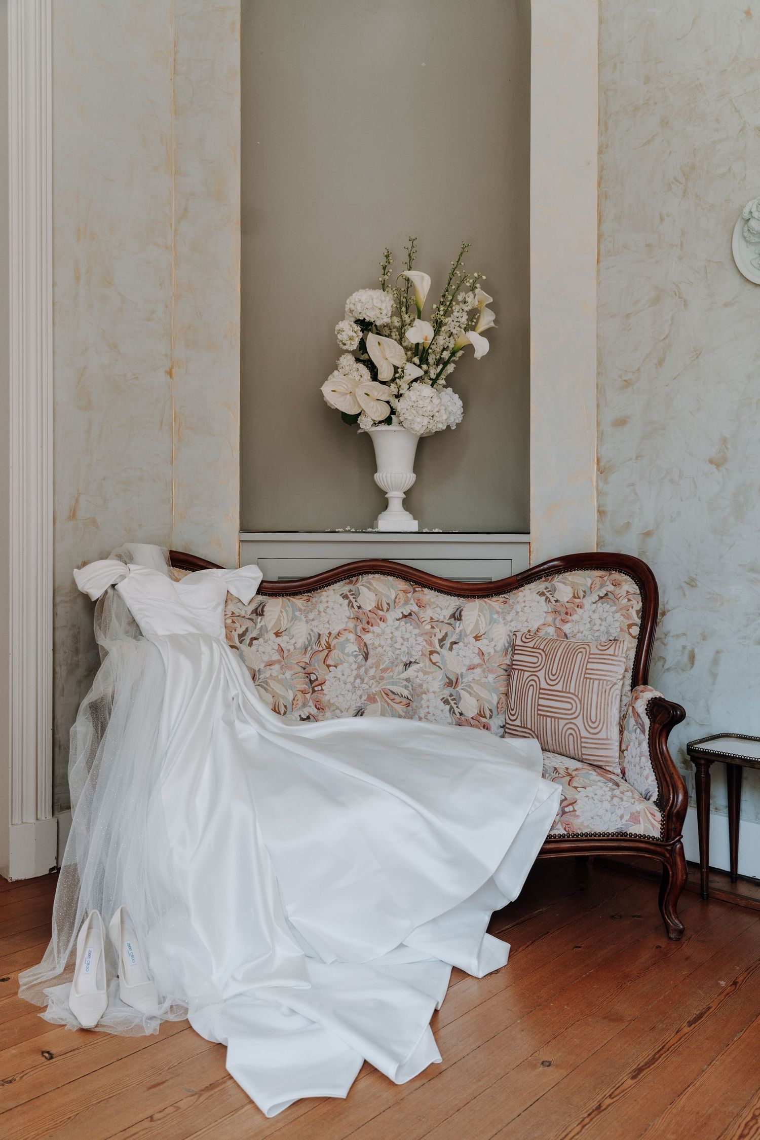 White wedding dress draped over vintage floral settee with flower arrangement.