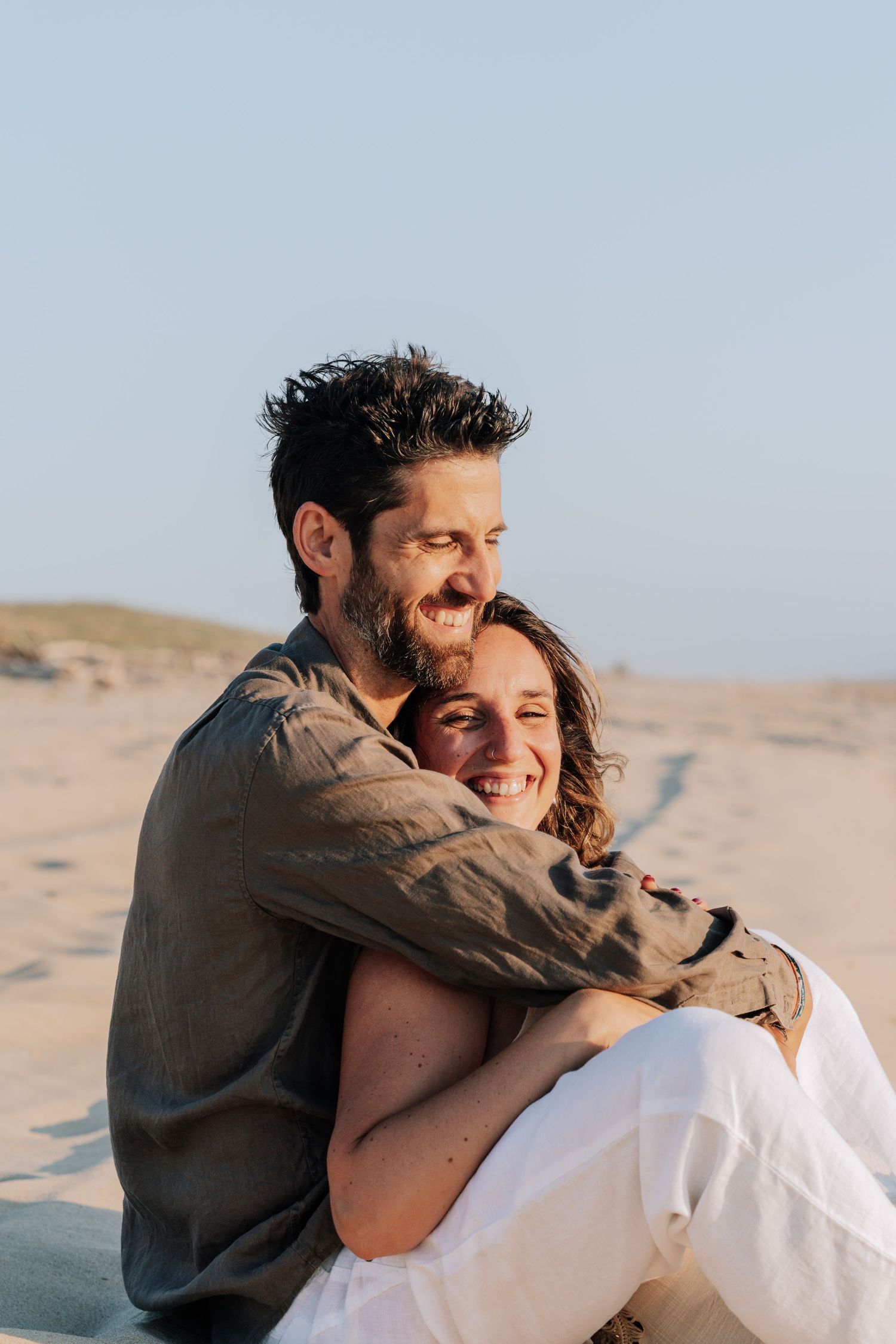 A couple embraces and laughs joyfully together while sitting on sandy dunes at sunset.