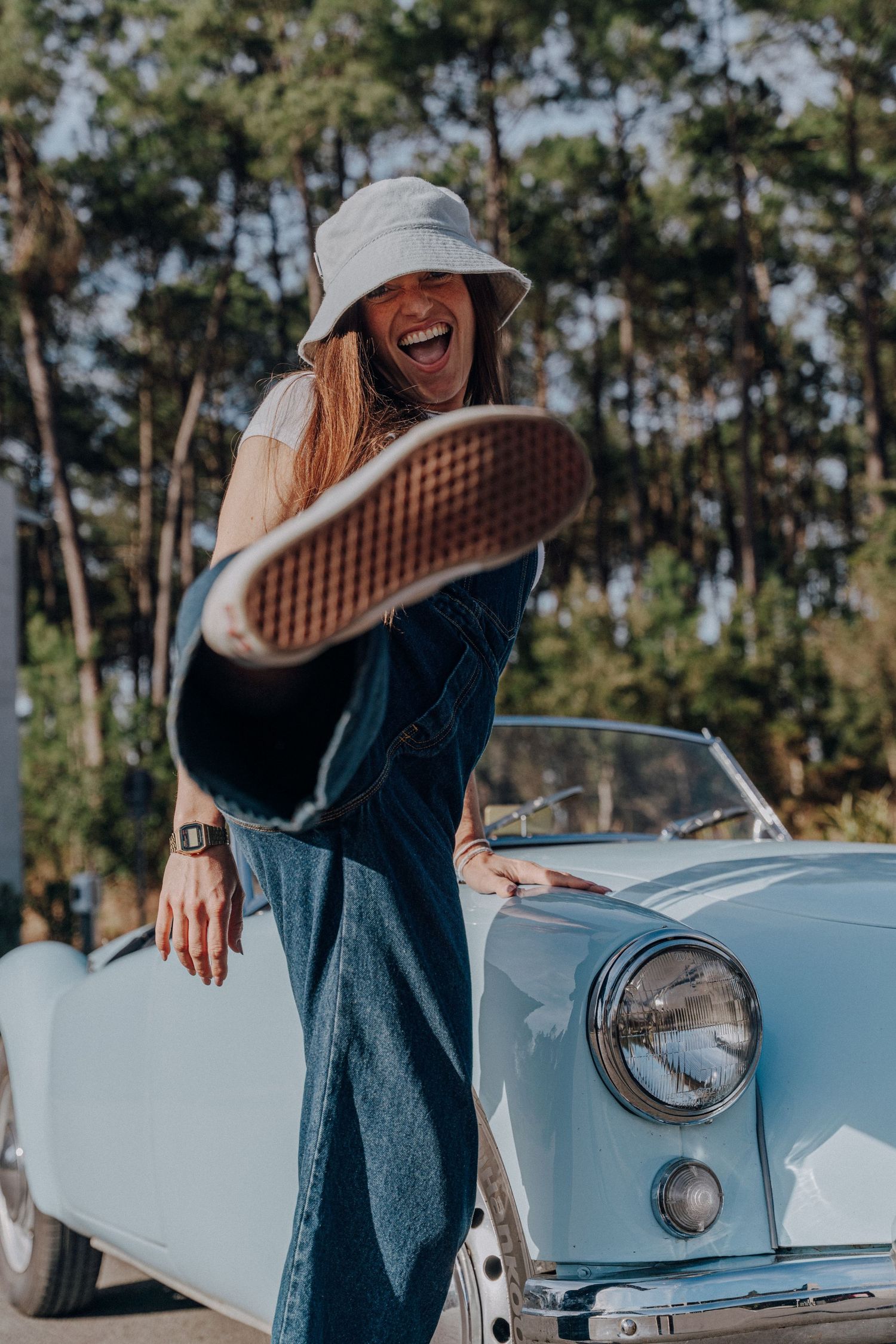 Person in denim and sun hat poses playfully with light blue classic car outdoors.