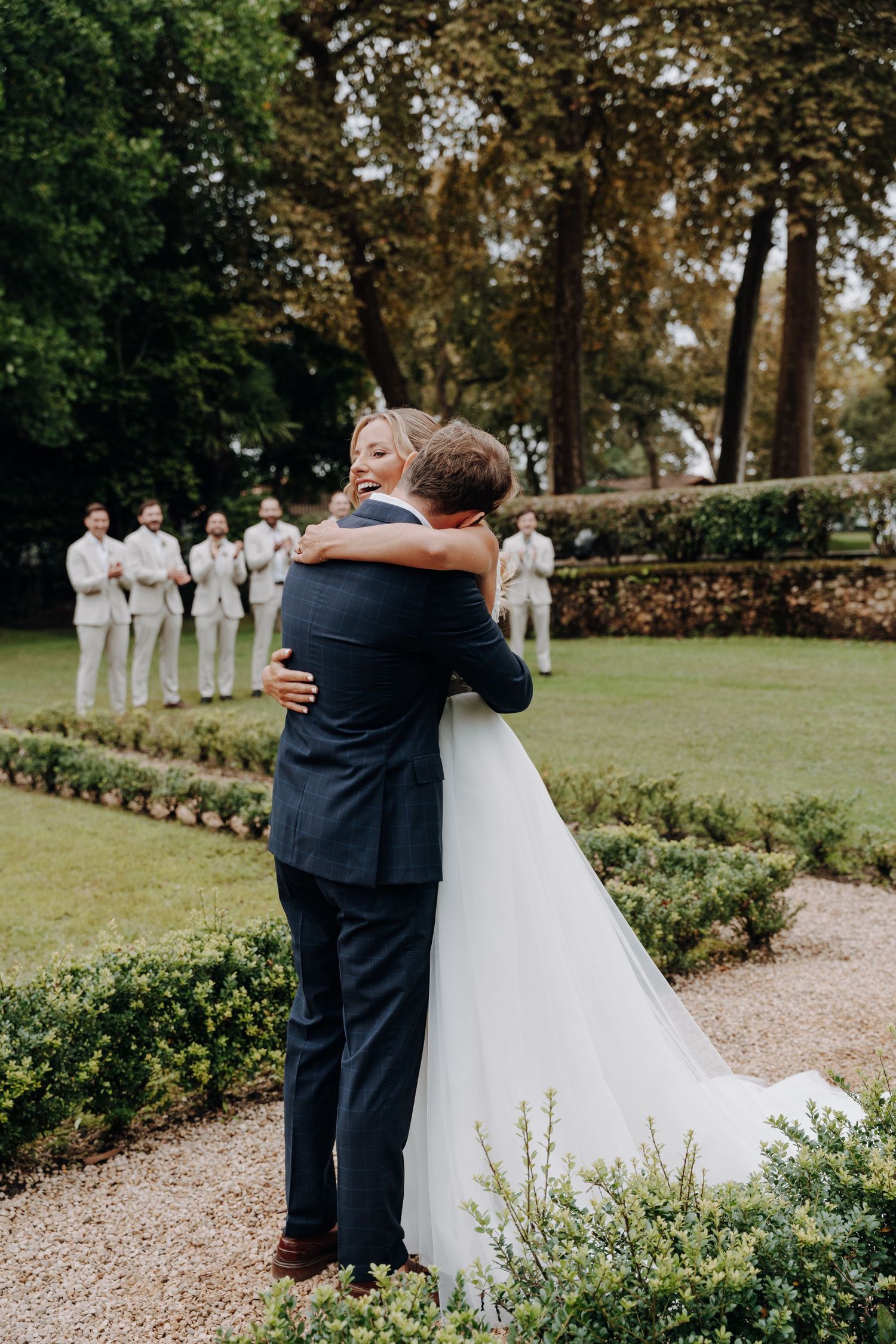 Wedding couple embraces in garden while groomsmen stand in background.