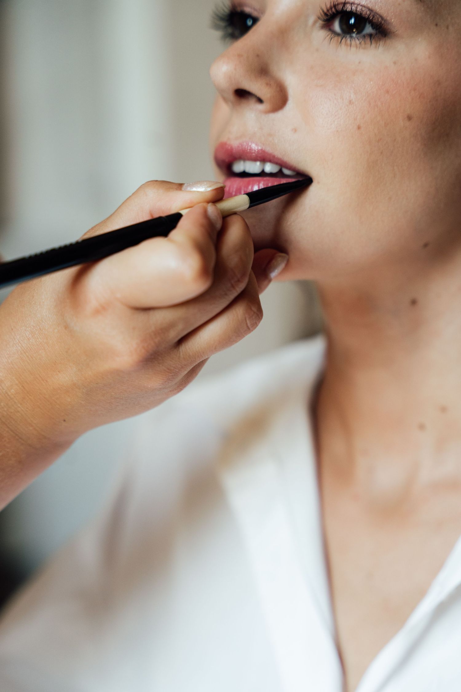 Makeup artist applying lip liner to bride with freckles wearing white robe before wedding ceremony.