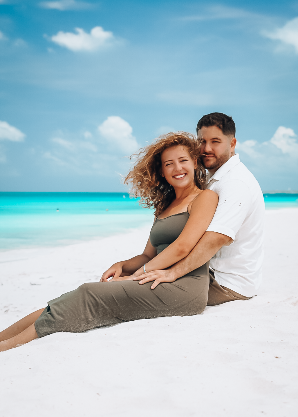 Couple sitting together on white sand beach with turquoise ocean, woman smiling in gray dress, man in white shirt embracing her.